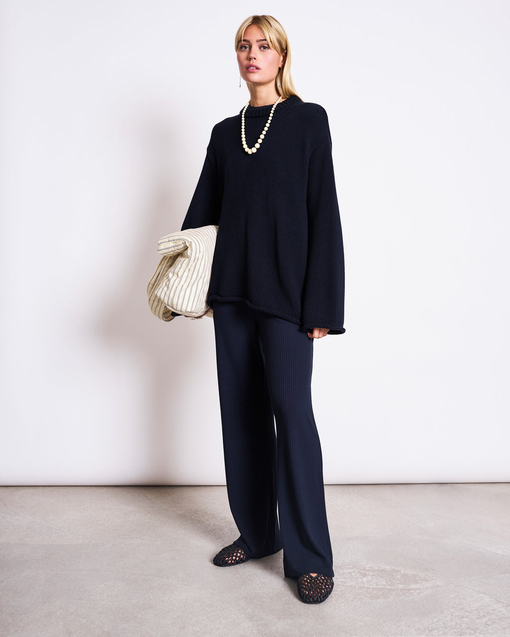 A person stands against a white wall wearing the jan-n-june JUMPER SONA NIGHTFALL GOTS, oversized black pants, black shoes, a chunky pearl necklace, and holding a large cream-colored bag under their arm.