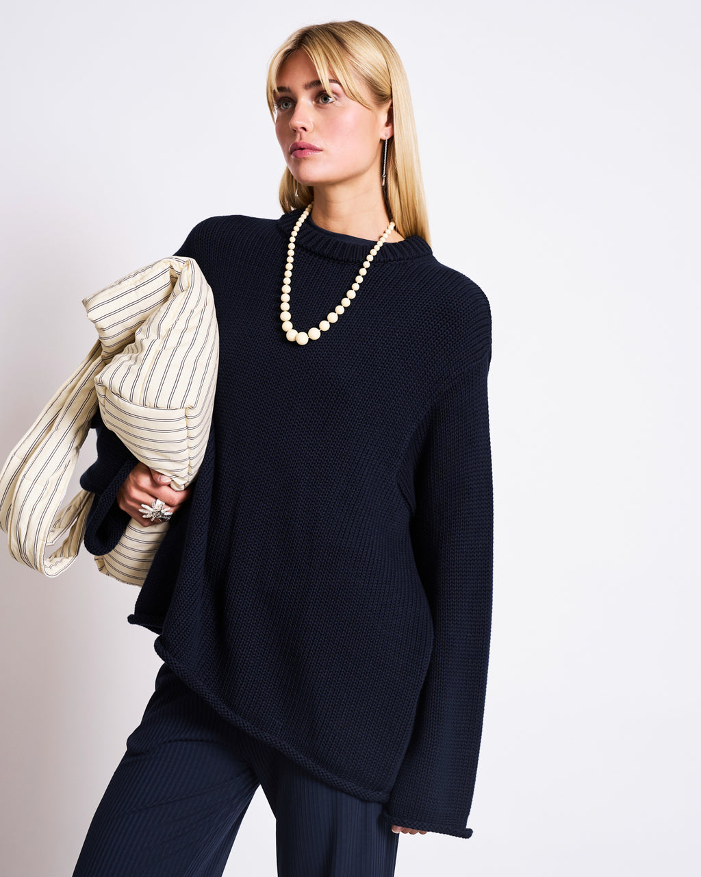 A woman with straight blonde hair wears the jan-n-june JUMPER SONA NIGHTFALL GOTS, an oversized organic cotton knit sweater, paired with dark pants and a beaded necklace, holding a striped blazer over her shoulder against a white background.