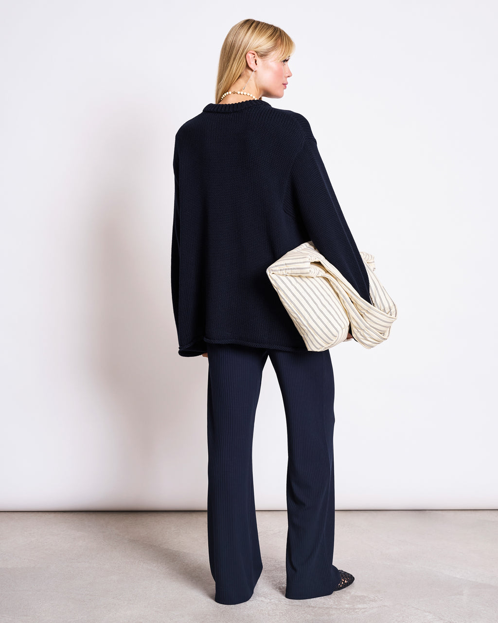 A woman with blonde hair faces away, wearing the jan-n-june JUMPER SONA NIGHTFALL GOTS—an oversized black sweater made from GOTS certified organic cotton—with matching pants and a large cream quilted bag, set against a plain white background.