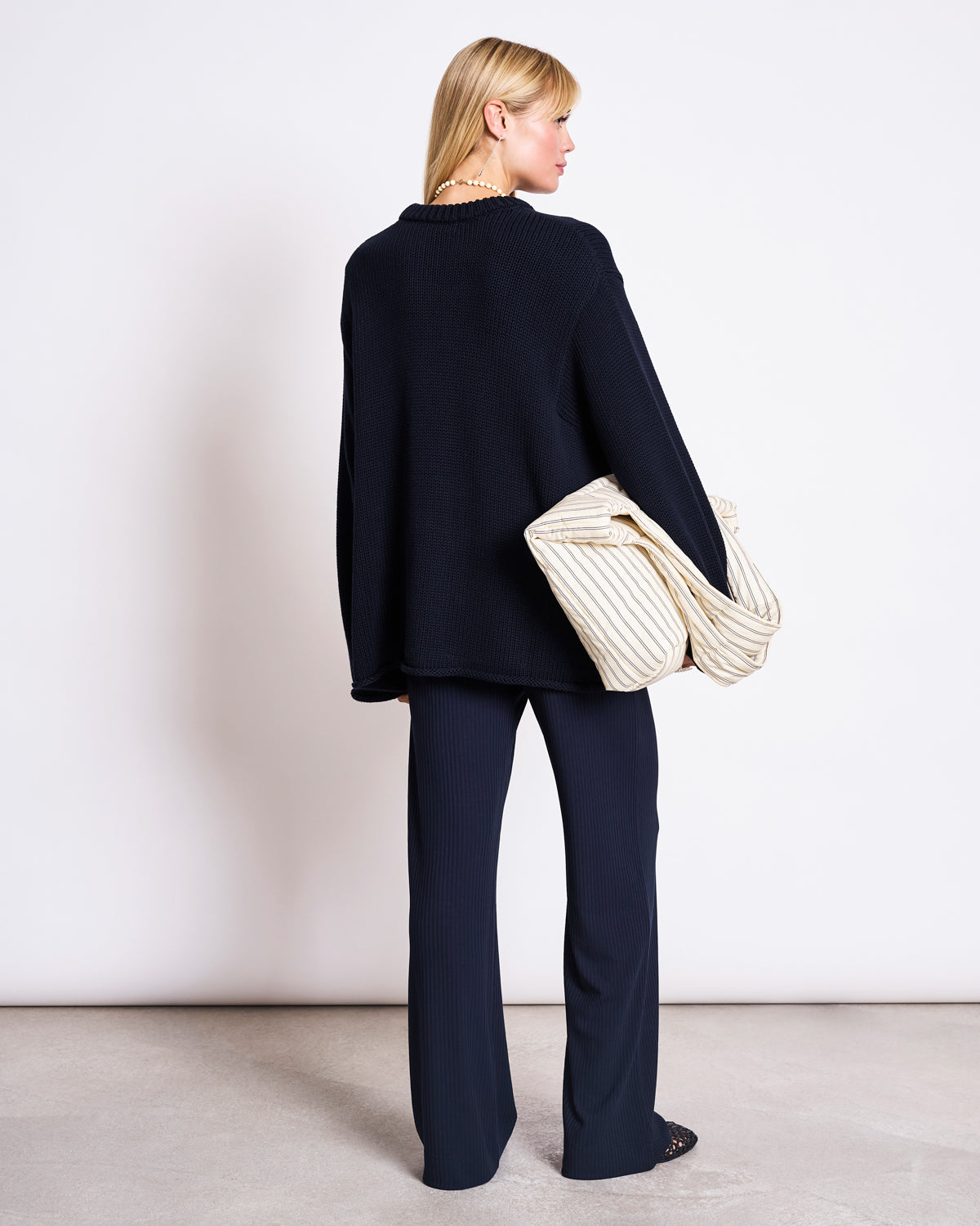 A woman with blonde hair faces away, wearing the jan-n-june JUMPER SONA NIGHTFALL GOTS—an oversized black sweater made from GOTS certified organic cotton—with matching pants and a large cream quilted bag, set against a plain white background.