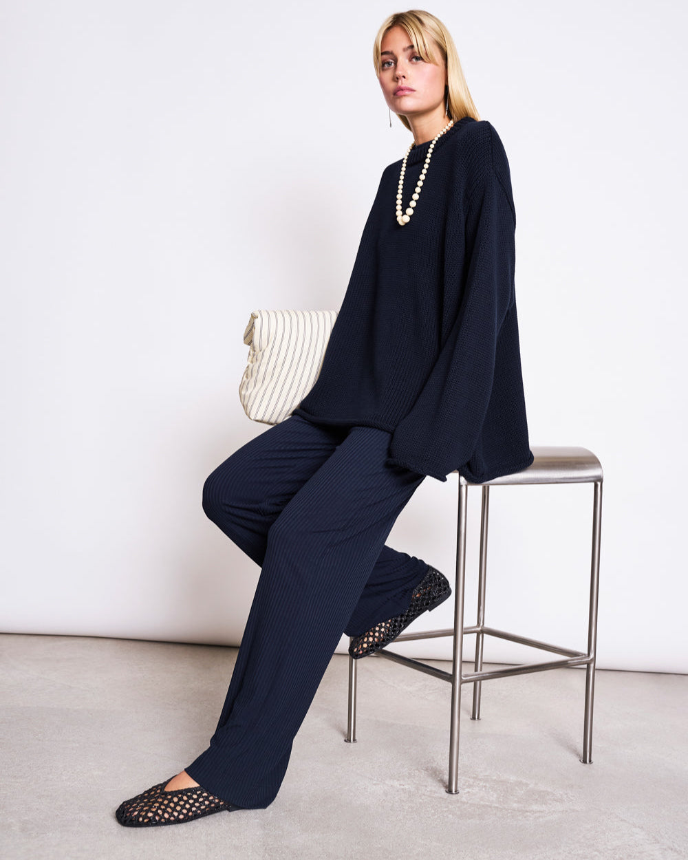 A person with blond hair sits on a tall metal stool, wearing the jan-n-june JUMPER SONA NIGHTFALL GOTS—an oversized black knit made from GOTS certified organic cotton—paired with black mesh flats and a white textured clutch, against a plain light background.