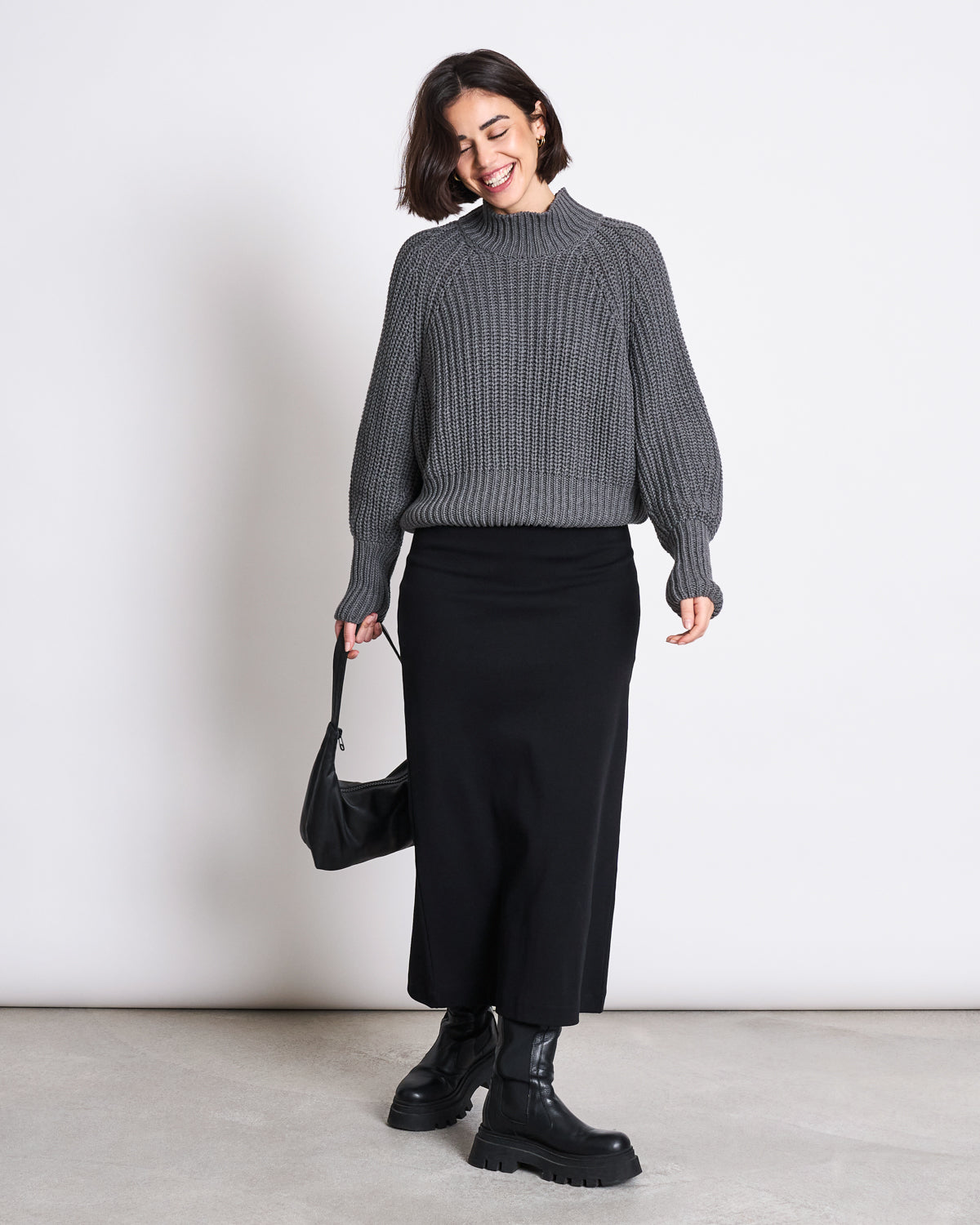A woman smiles in a chunky gray knit sweater, the jan-n-june MAXI SKIRT BURNITA BLACK GOTS, and black chunky boots. She holds a small black handbag and poses against a plain white background.