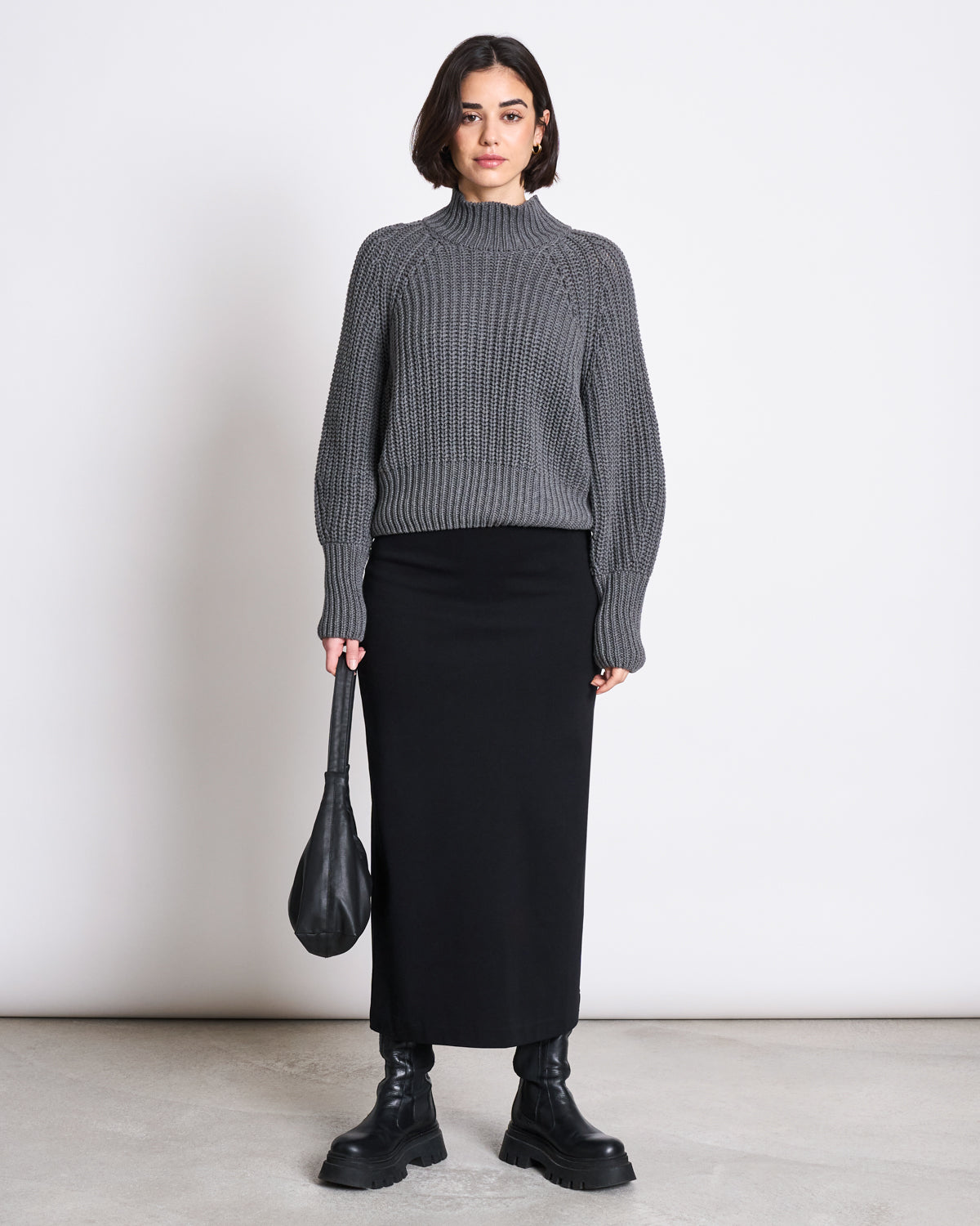 A woman with short dark hair and a neutral expression wears the jan-n-june MAXI SKIRT BURNITA BLACK GOTS, a gray textured sweater, chunky boots, and holds a black bag. The elegant maxi skirt complements her minimalist style.