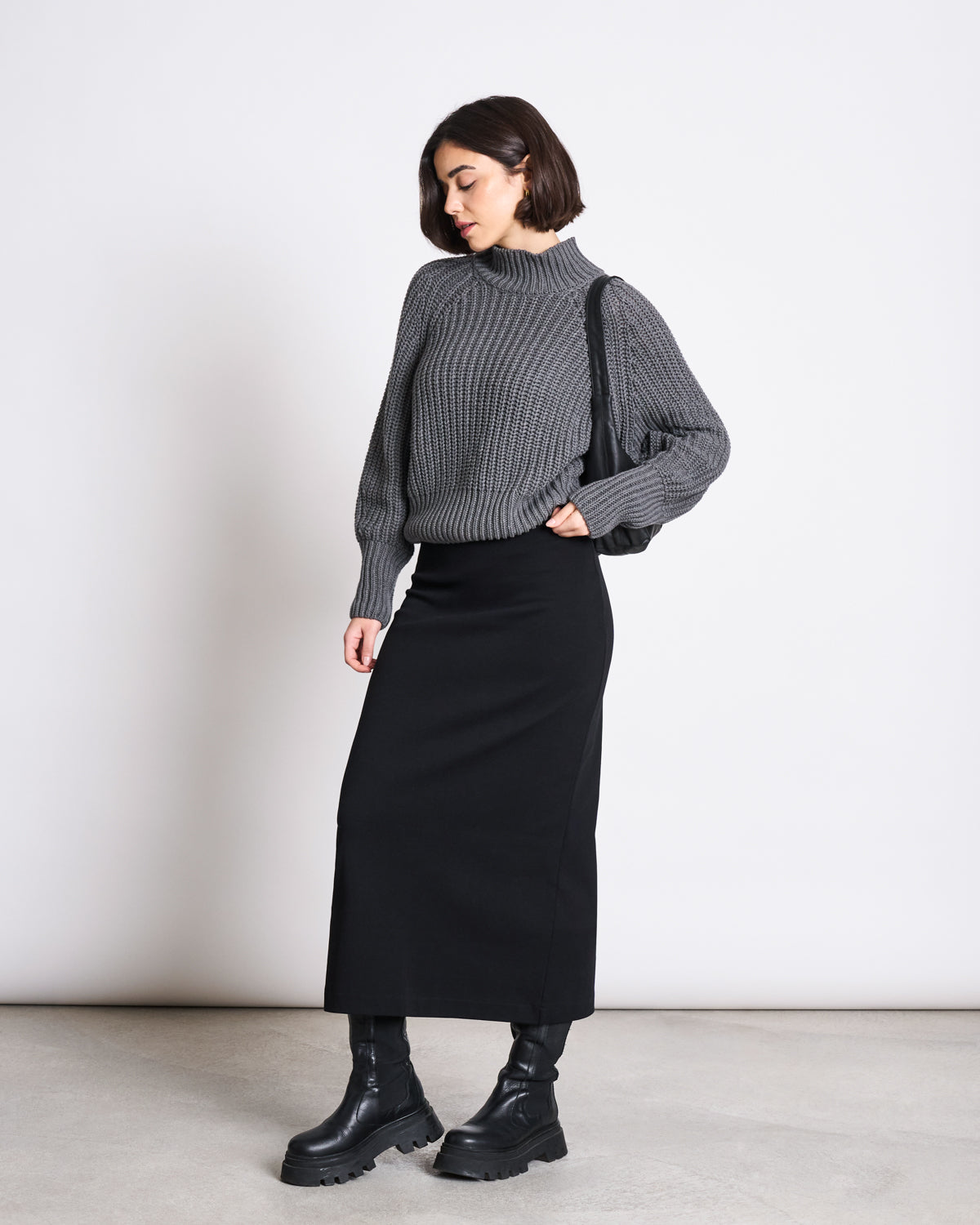 A woman stands against a plain background wearing the jan-n-june MAXI SKIRT BURNITA BLACK GOTS, a gray knit sweater, chunky black boots, and a black shoulder bag. She looks down with one hand in her pocket.