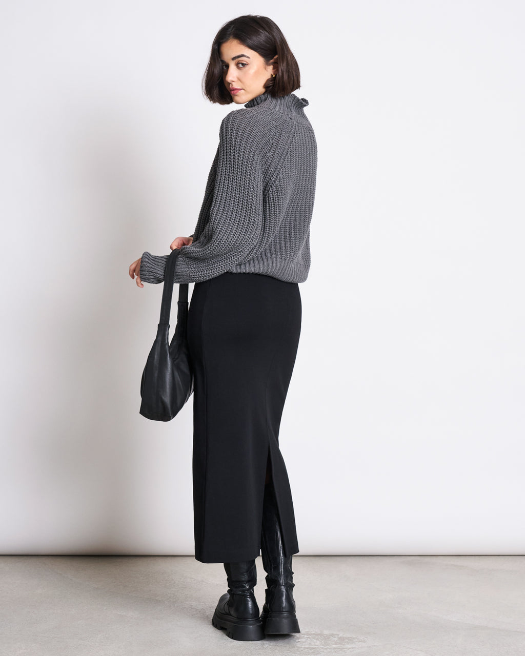 A woman with a bob haircut wears a gray knit sweater, jan-n-junes MAXI SKIRT BURNITA BLACK GOTS, black boots, and carries a black shoulder bag as she stands facing away but looks back over her shoulder against a white background.