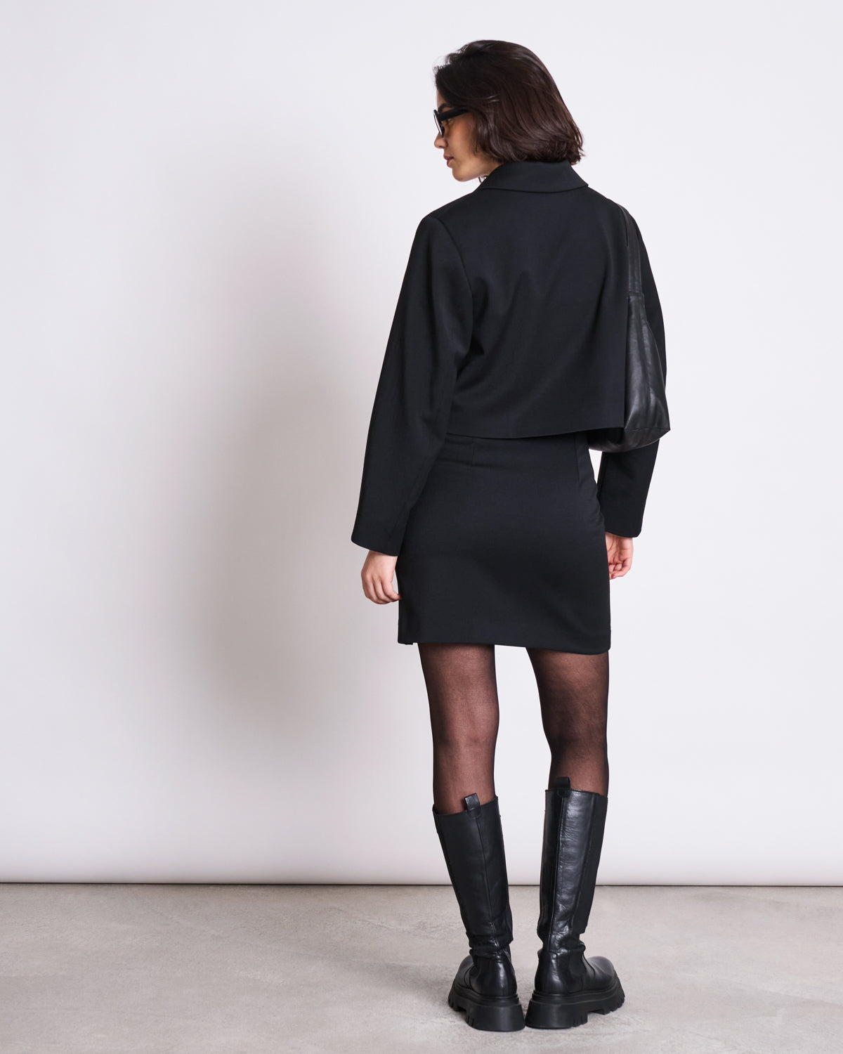 A woman with short dark hair and sunglasses faces away, wearing the jan-n-june MINI SKIRT AINOHA BLACK made from recycled polyester, a matching cropped jacket, sheer black tights, and knee-high boots against a plain light background.