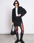 A woman wears AINOHA sunglasses, a cropped black blazer, white top, MINI SKIRT AINOHA BLACK by jan-n-june, sheer tights, knee-high boots, and holds a recycled polyester handbag against a plain background.