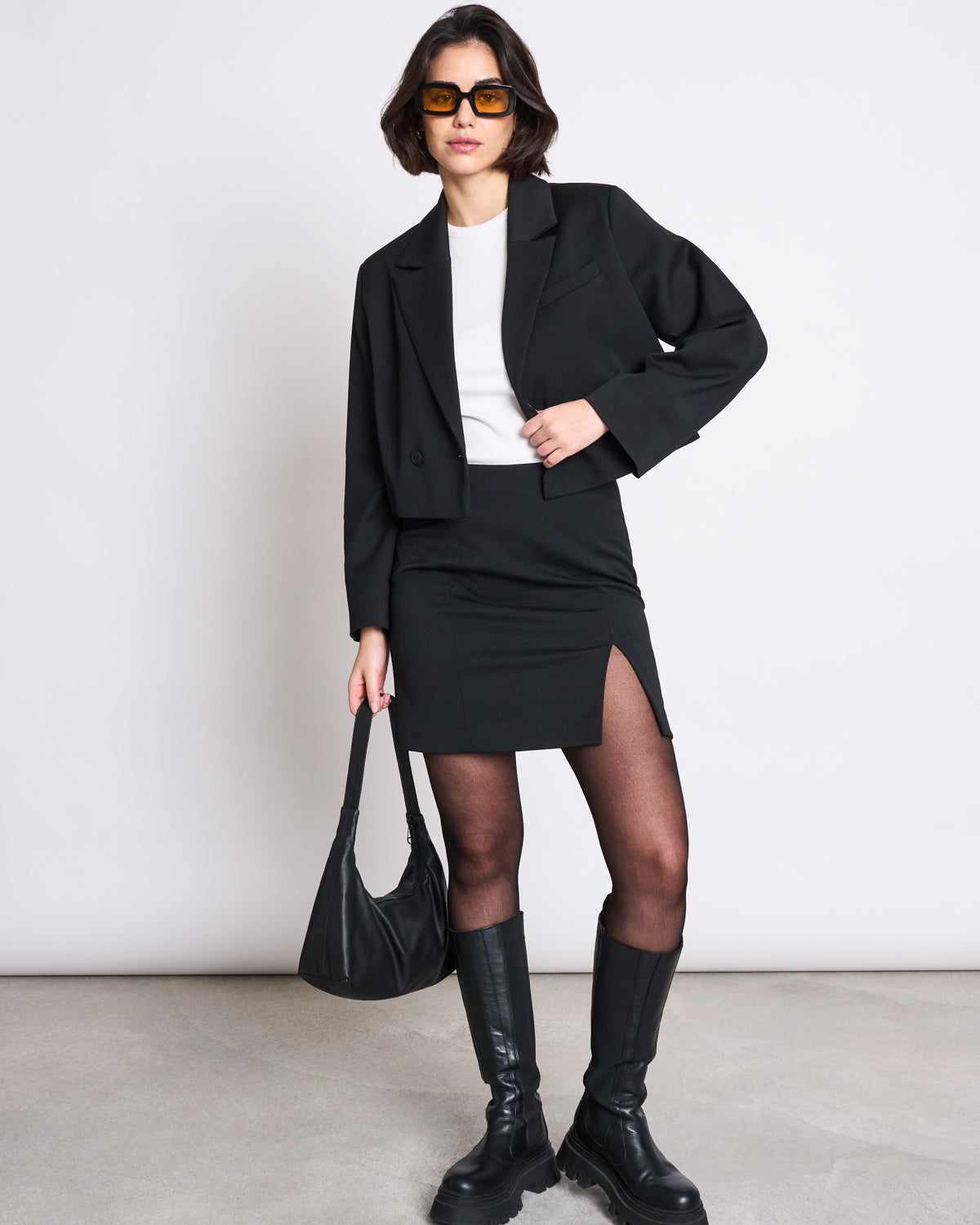 A woman wears AINOHA sunglasses, a cropped black blazer, white top, MINI SKIRT AINOHA BLACK by jan-n-june, sheer tights, knee-high boots, and holds a recycled polyester handbag against a plain background.