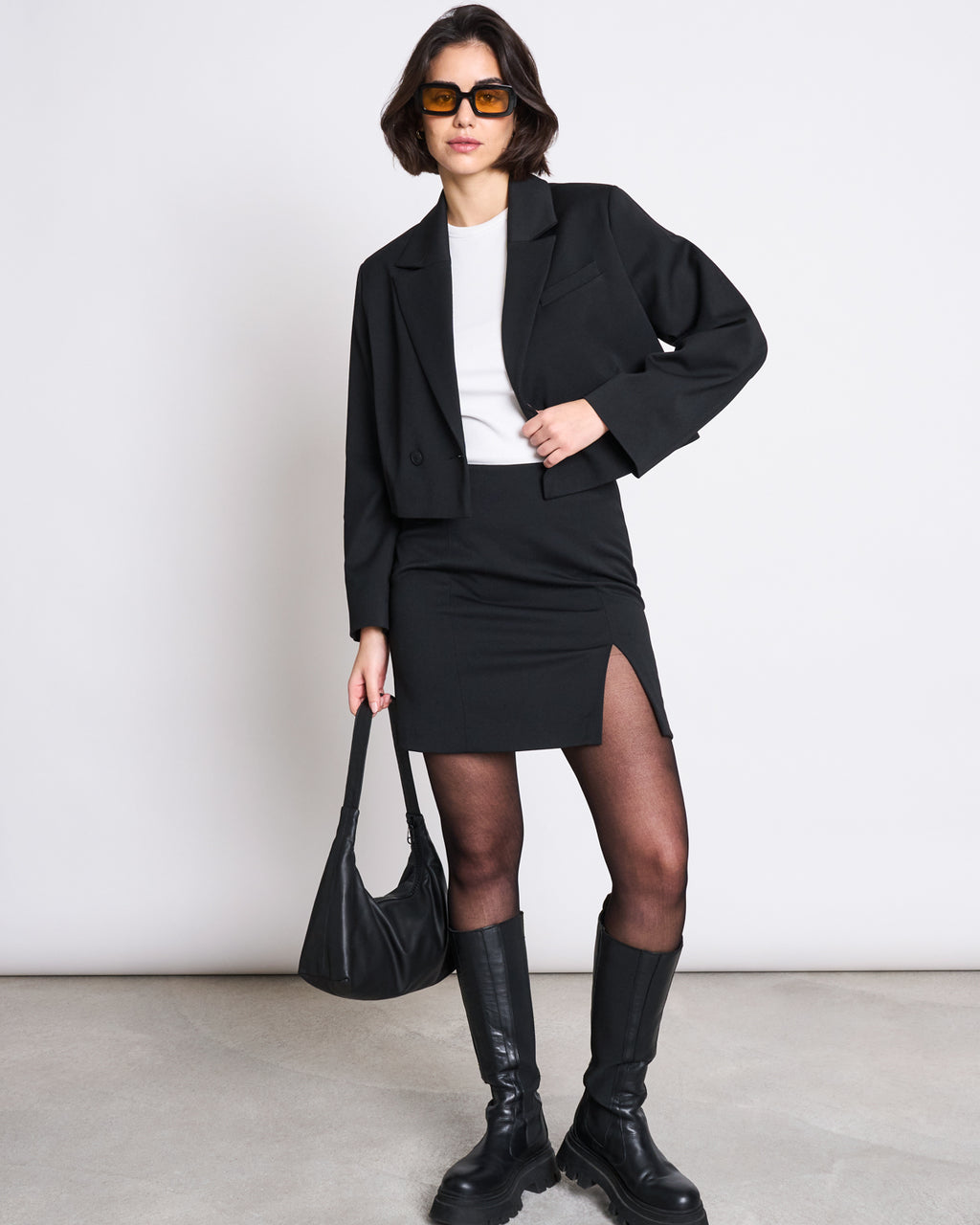 A woman wearing the jan-n-june CROPPED BLAZER TULIA BLACK, oversized sunglasses, a white top, a black mini skirt with a front slit, black tights, knee-high boots, and holding a black shoulder bag stands against a plain background.