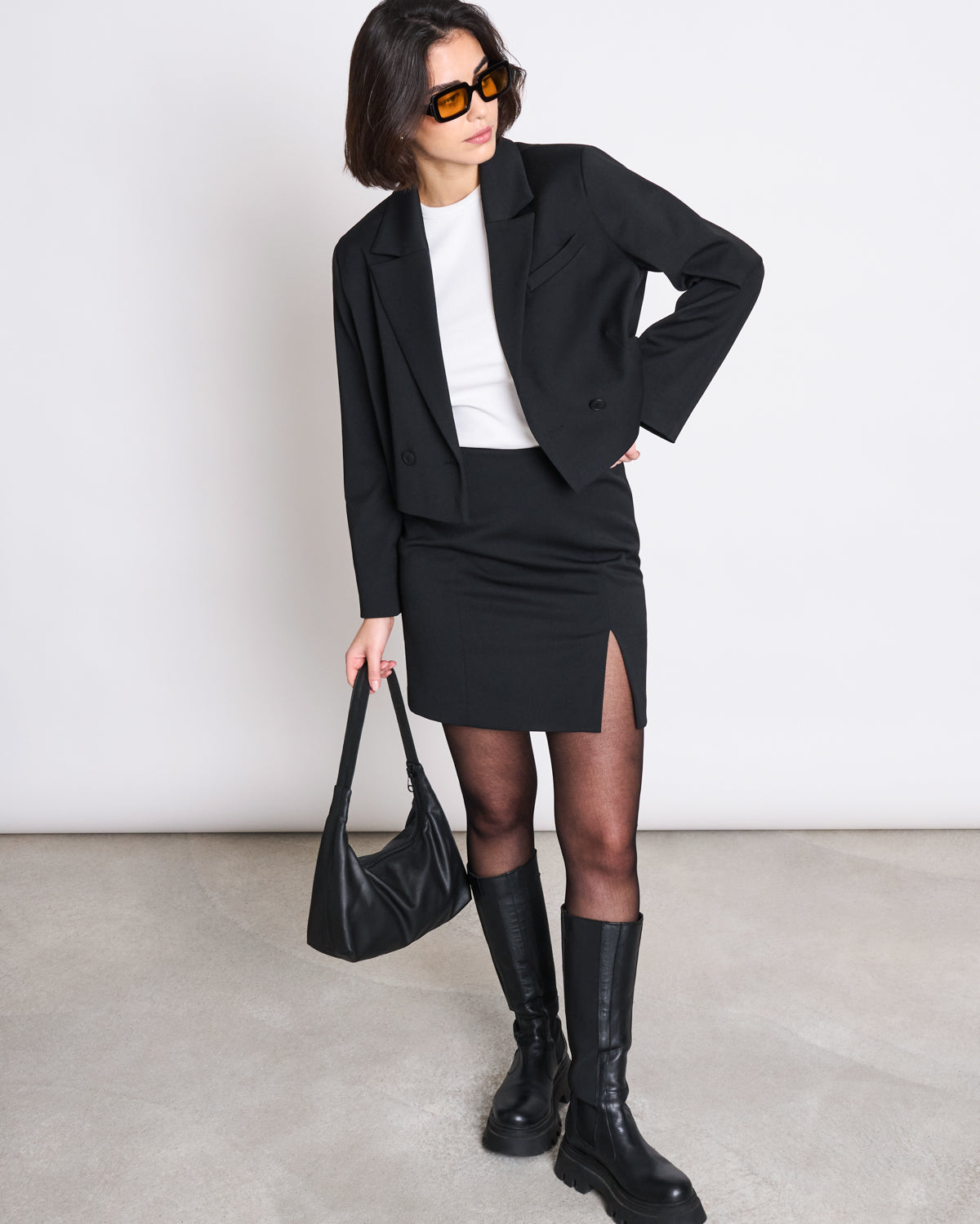 A woman stylishly poses against a plain light background wearing the jan-n-june CROPPED BLAZER TULIA BLACK over a white top, paired with a black mini skirt, sheer tights, knee-high boots, sunglasses, and holding a black handbag.