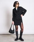A woman wears the jan-n-june CROPPED BLAZER TULIA BLACK with a black skirt, tights, chunky boots, sunglasses, and holds a black handbag, posing against a plain light background.