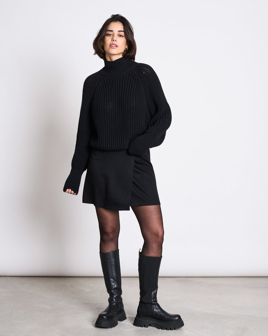 A person with short dark hair and a neutral expression stands against a plain white wall wearing the jan-n-june JUMPER OLA BLACK GOTS, paired with a black mini skirt, black tights, and black knee-high chunky boots.