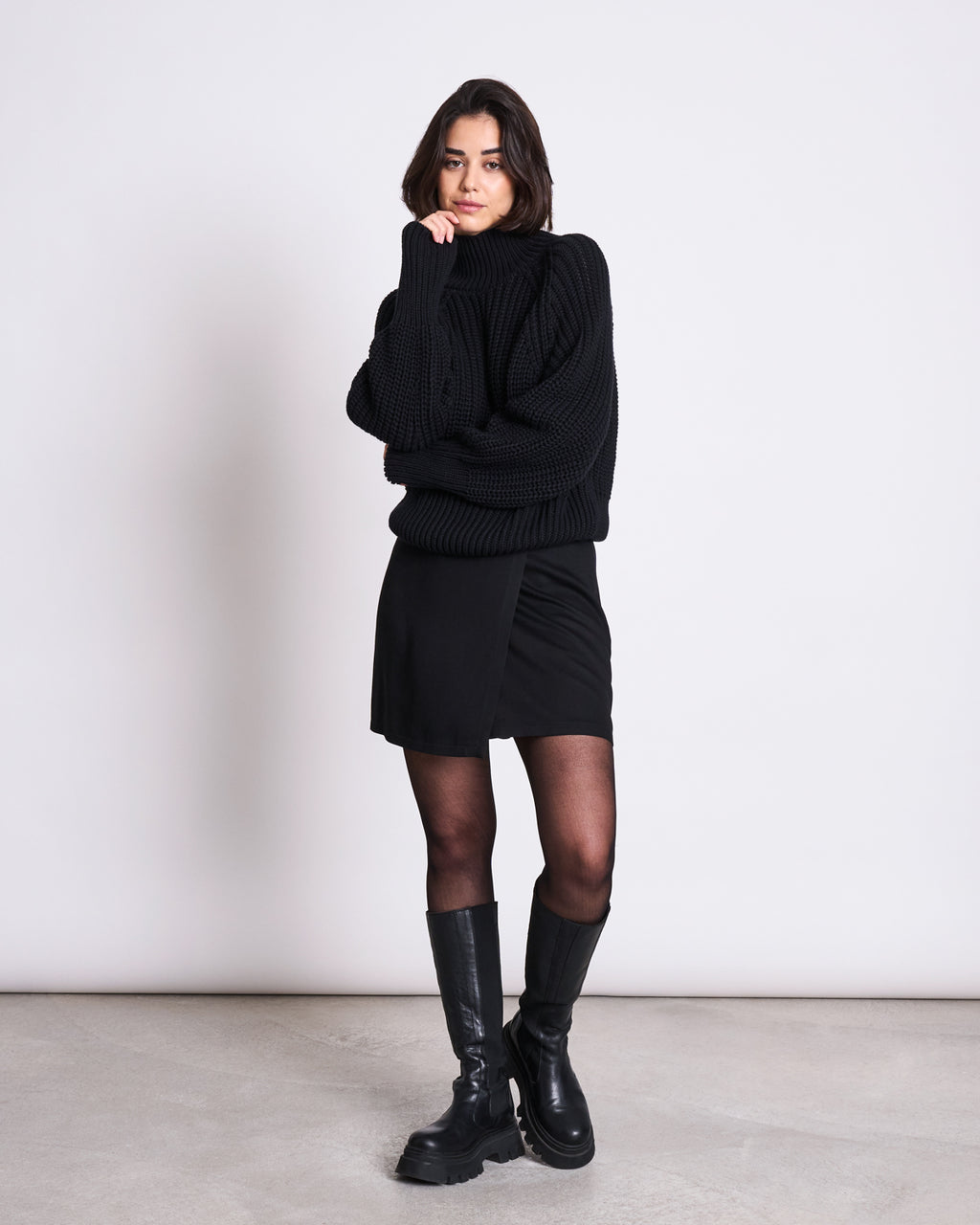 A woman with shoulder-length brown hair poses confidently against a plain light background, wearing the jan-n-june JUMPER OLA BLACK GOTS with a black mini skirt, tights, and knee-high boots, resting her chin on her hand.