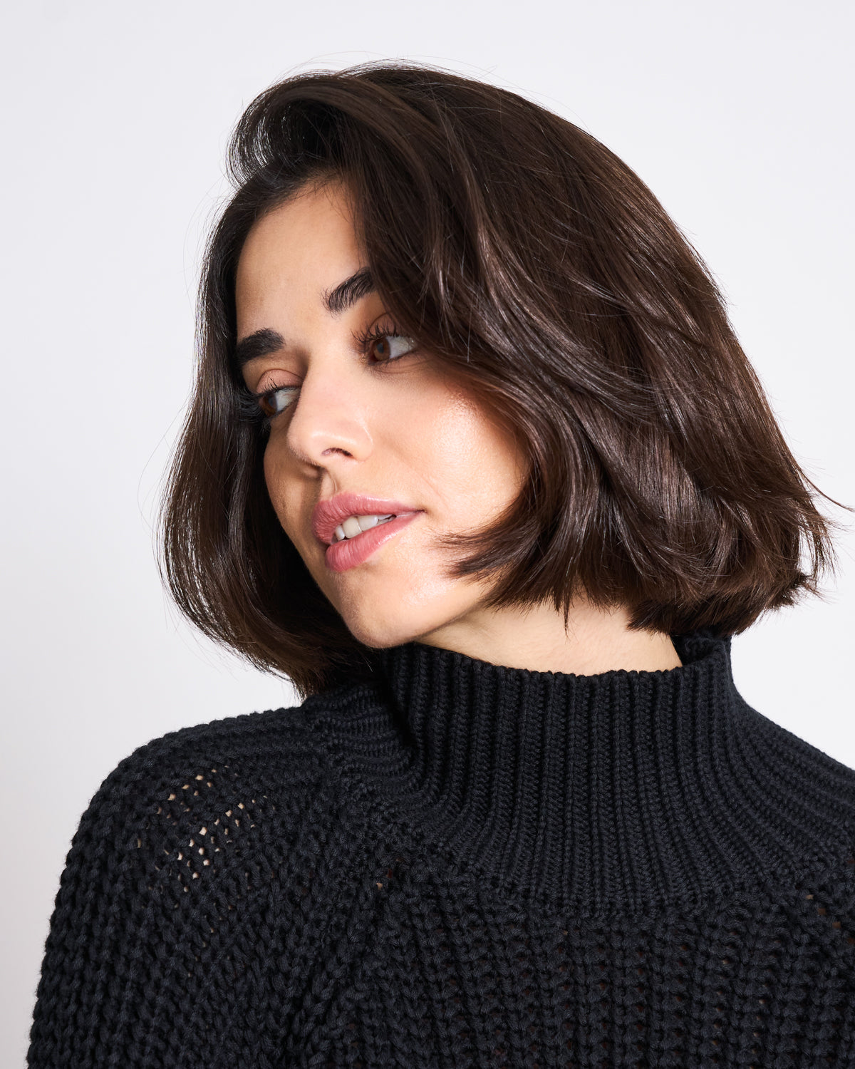 A woman with chin-length dark brown hair wears the jan-n-june JUMPER OLA BLACK GOTS, a chunky black turtleneck, and looks to the side with a neutral expression against a plain white background.