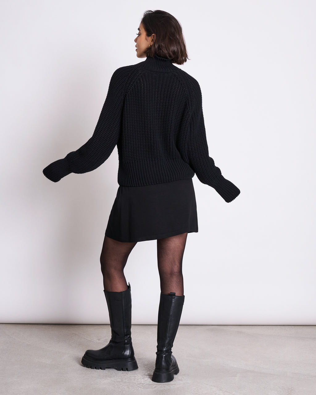 A woman faces away from the camera, wearing the jan-n-june JUMPER OLA BLACK GOTS, a chunky turtleneck jumper, styled with a black mini skirt, sheer black tights, and knee-high boots against a plain white background.