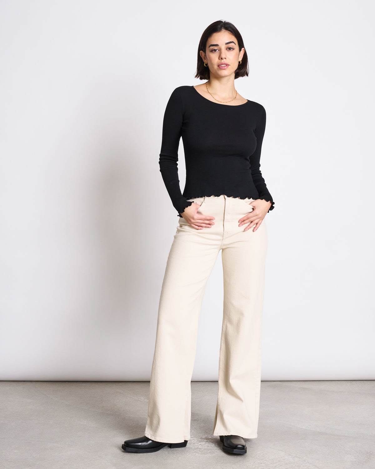A woman with short dark hair and a neutral expression stands against a white wall, wearing the jan-n-june MINIRIB LONGSLEEVE AVERSA BLACK GOTS top, beige high-waisted wide-leg pants, and black shoes.