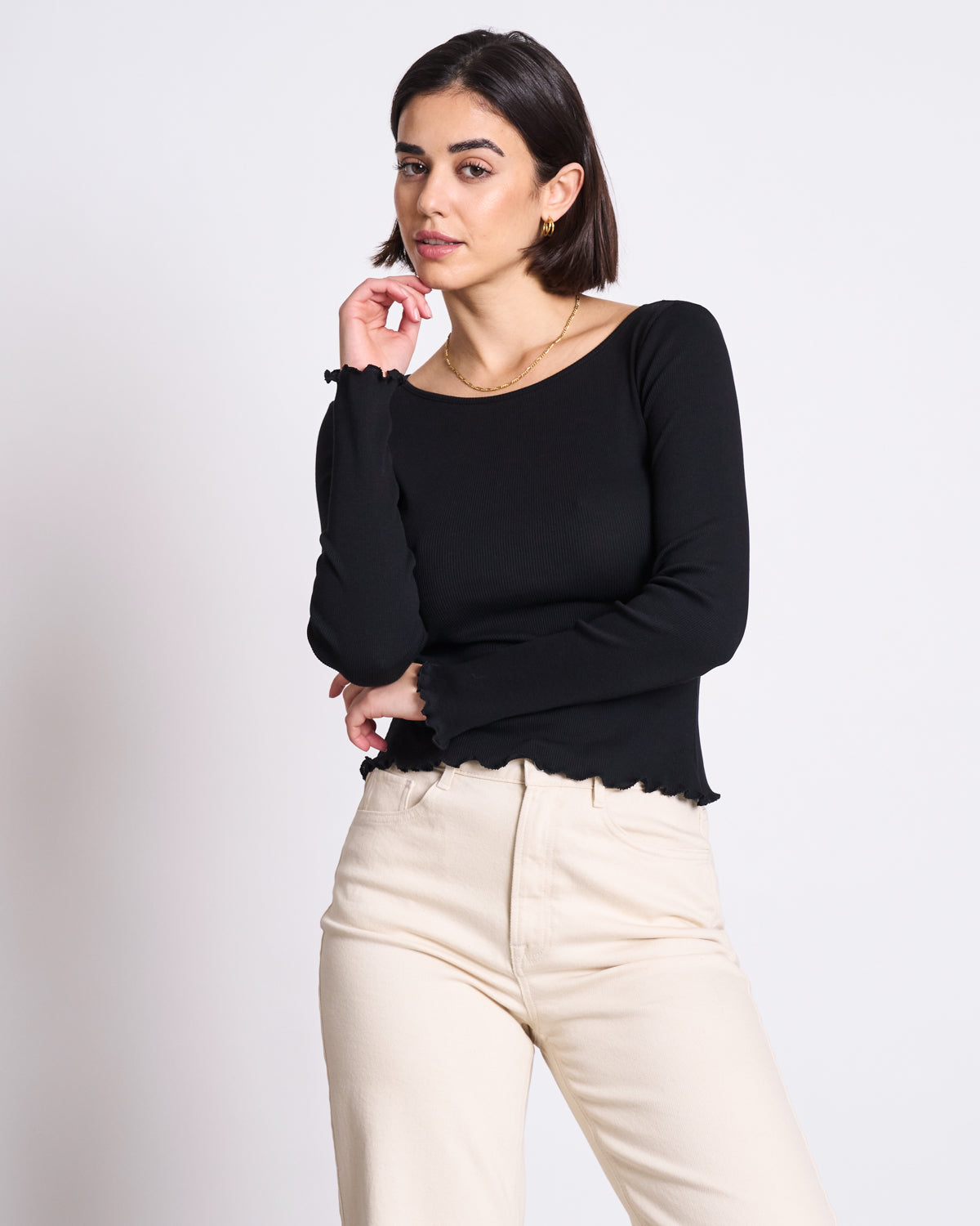 A woman with short dark hair wears the jan-n-june MINIRIB LONGSLEEVE AVERSA BLACK GOTS crop top and high-waisted beige pants, standing against a white background and gazing at the camera with one hand touching her chin.