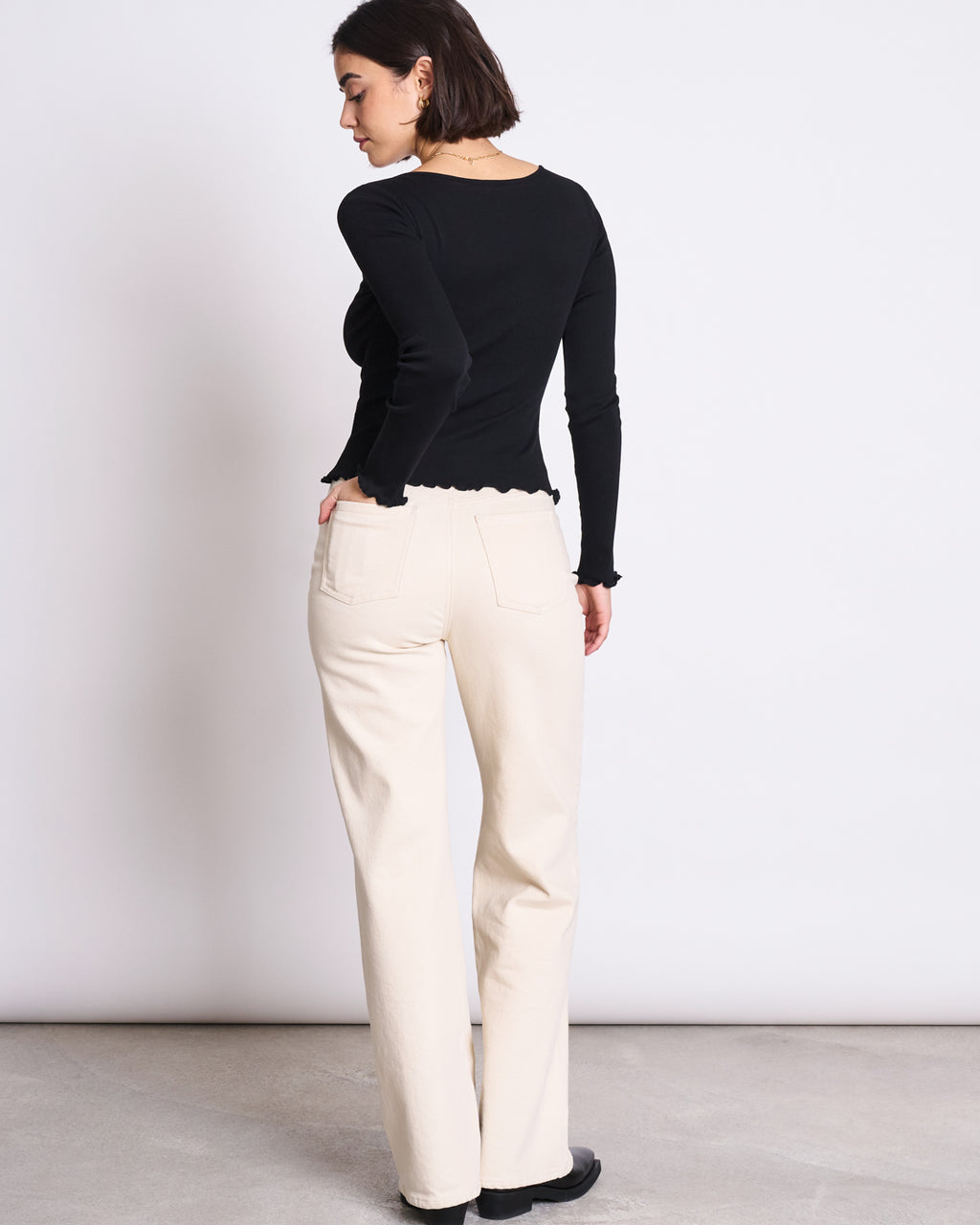 A woman with short dark hair stands with her back to the camera, wearing jan-n-junes MINIRIB LONGSLEEVE AVERSA BLACK GOTS top and high-waisted cream organic cotton pants, paired with black shoes, set against a plain white background.