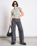 A woman in a jan-n-june MINIRIB T-SHIRT REN MUTED SEA SPRAY GOTS, loose gray scallop-hem jeans, black chunky shoes, and sunglasses holds a black handbag at her side against a white background.