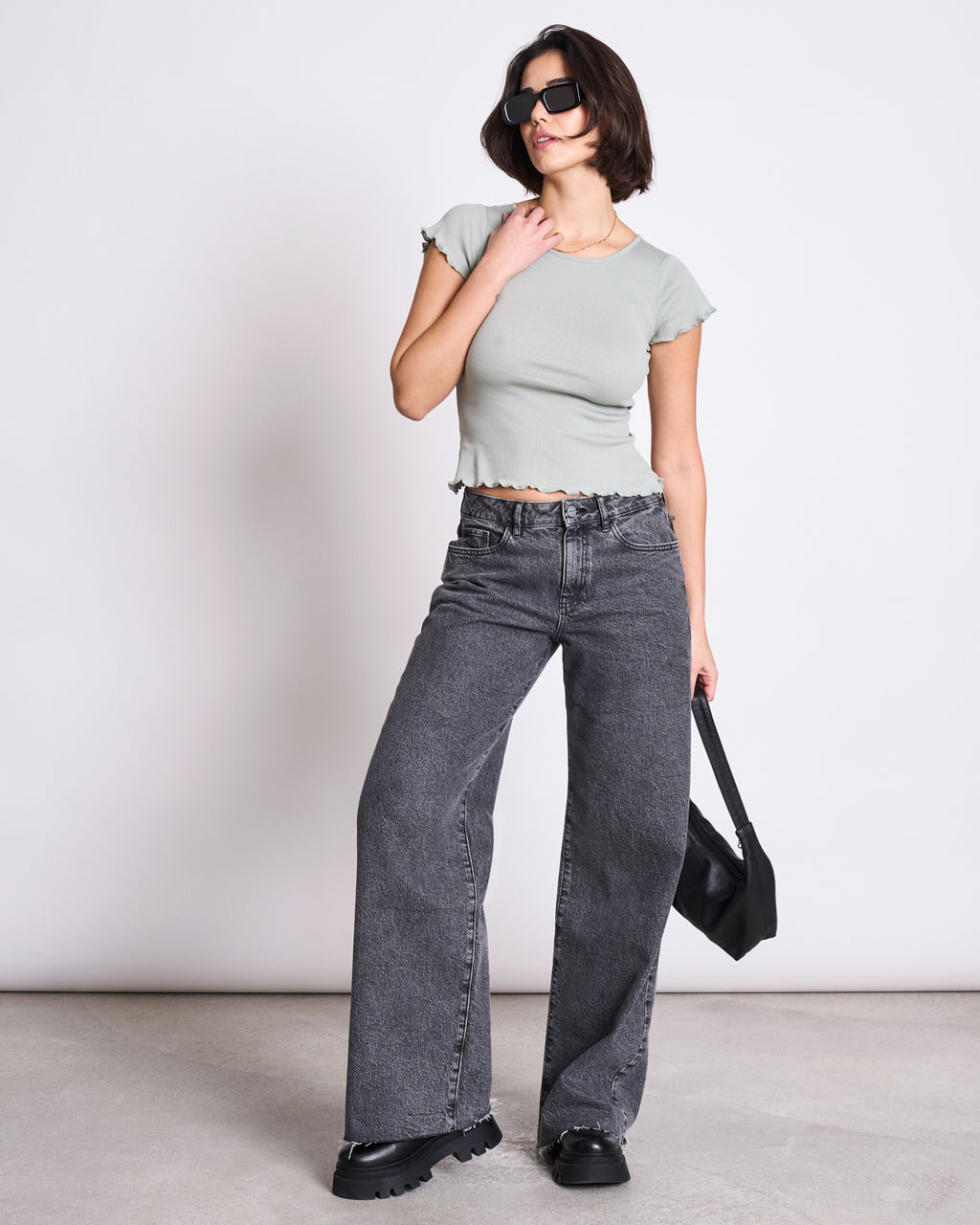 A woman stands against a plain wall wearing the jan-n-june MINIRIB T-SHIRT REN MUTED SEA SPRAY GOTS, high-waisted wide-leg black jeans, chunky black shoes, sunglasses, and holding a black handbag.