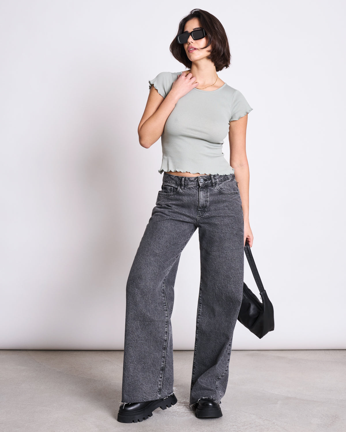 A woman stands against a plain wall wearing the jan-n-june MINIRIB T-SHIRT REN MUTED SEA SPRAY GOTS, high-waisted wide-leg black jeans, chunky black shoes, sunglasses, and holding a black handbag.
