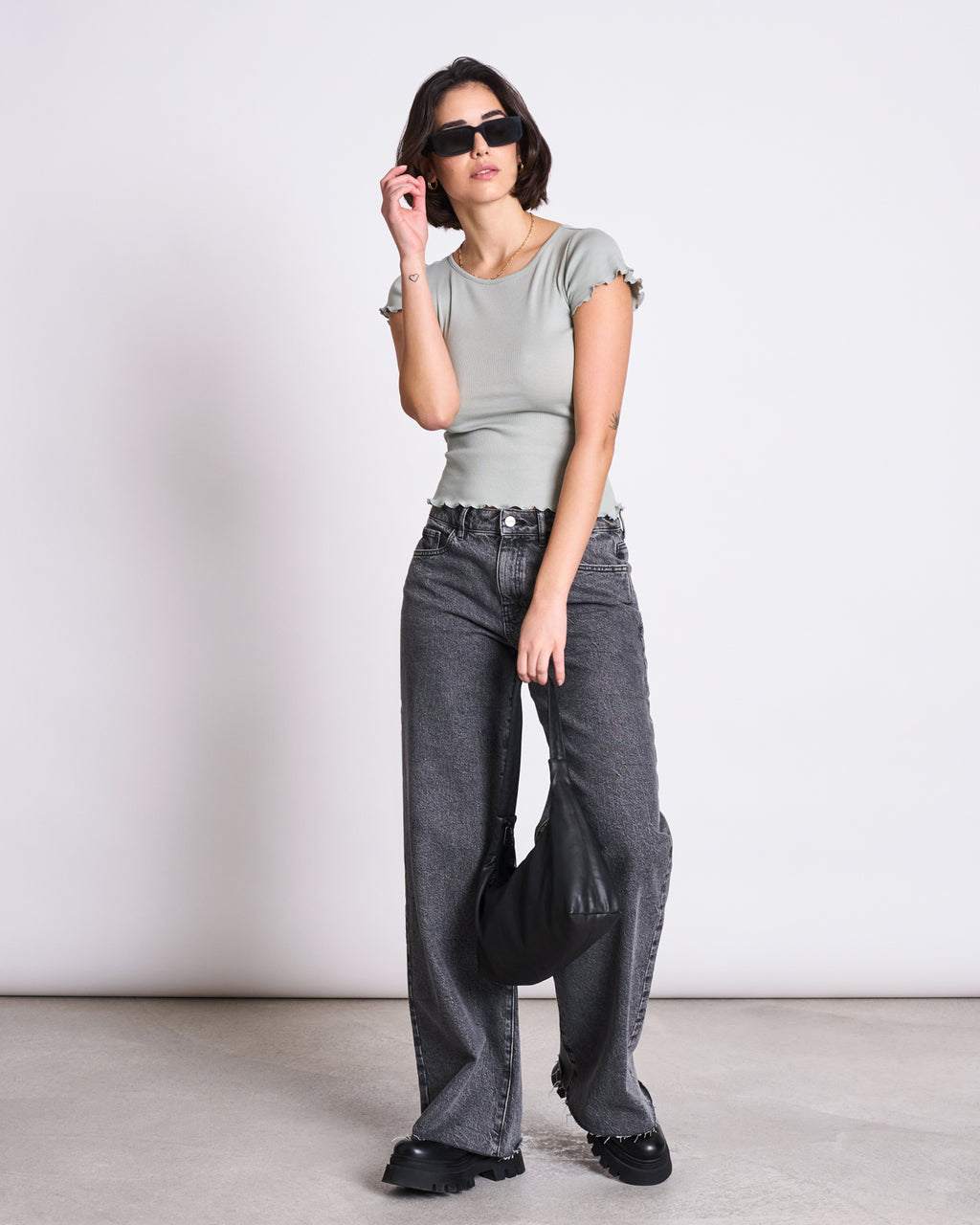 A woman stands against a plain background wearing jan-n-june’s MINIRIB T-SHIRT REN in Muted Sea Spray, wide-leg gray jeans with scalloped hems, sunglasses, black shoes, and holding a black handbag by her side.