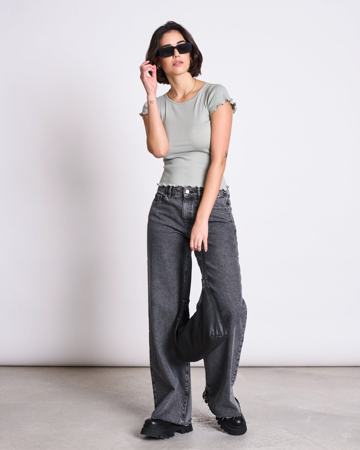 A woman stands against a plain background wearing jan-n-june’s MINIRIB T-SHIRT REN in Muted Sea Spray, wide-leg gray jeans with scalloped hems, sunglasses, black shoes, and holding a black handbag by her side.
