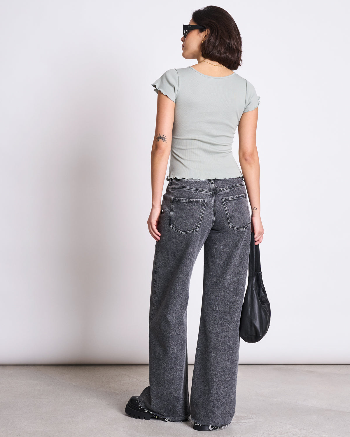 A woman with a short hairstyle and small arm tattoo faces away, wearing jan-n-junes MINIRIB T-SHIRT REN MUTED SEA SPRAY GOTS, loose high-waisted gray jeans, chunky black shoes, sunglasses, and holding a black bag.