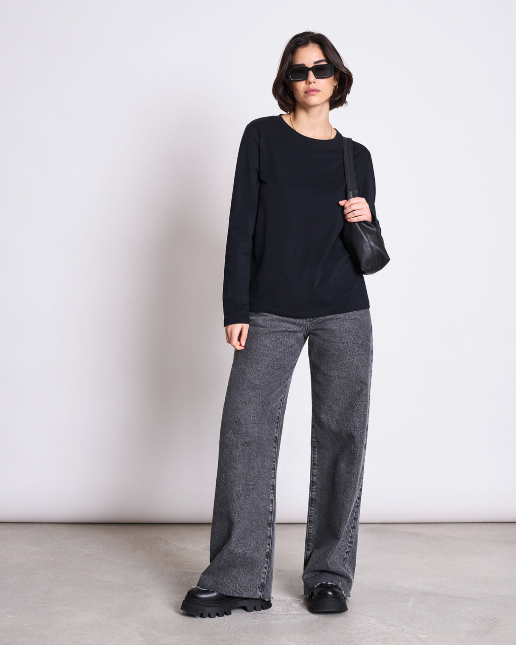 A woman stands against a plain, light background wearing jan-n-junes BRUSHED LONGSLEEVE BOY BLACK GOTS with black sunglasses, gray wide-leg jeans, chunky black shoes, and holding a small black shoulder bag.