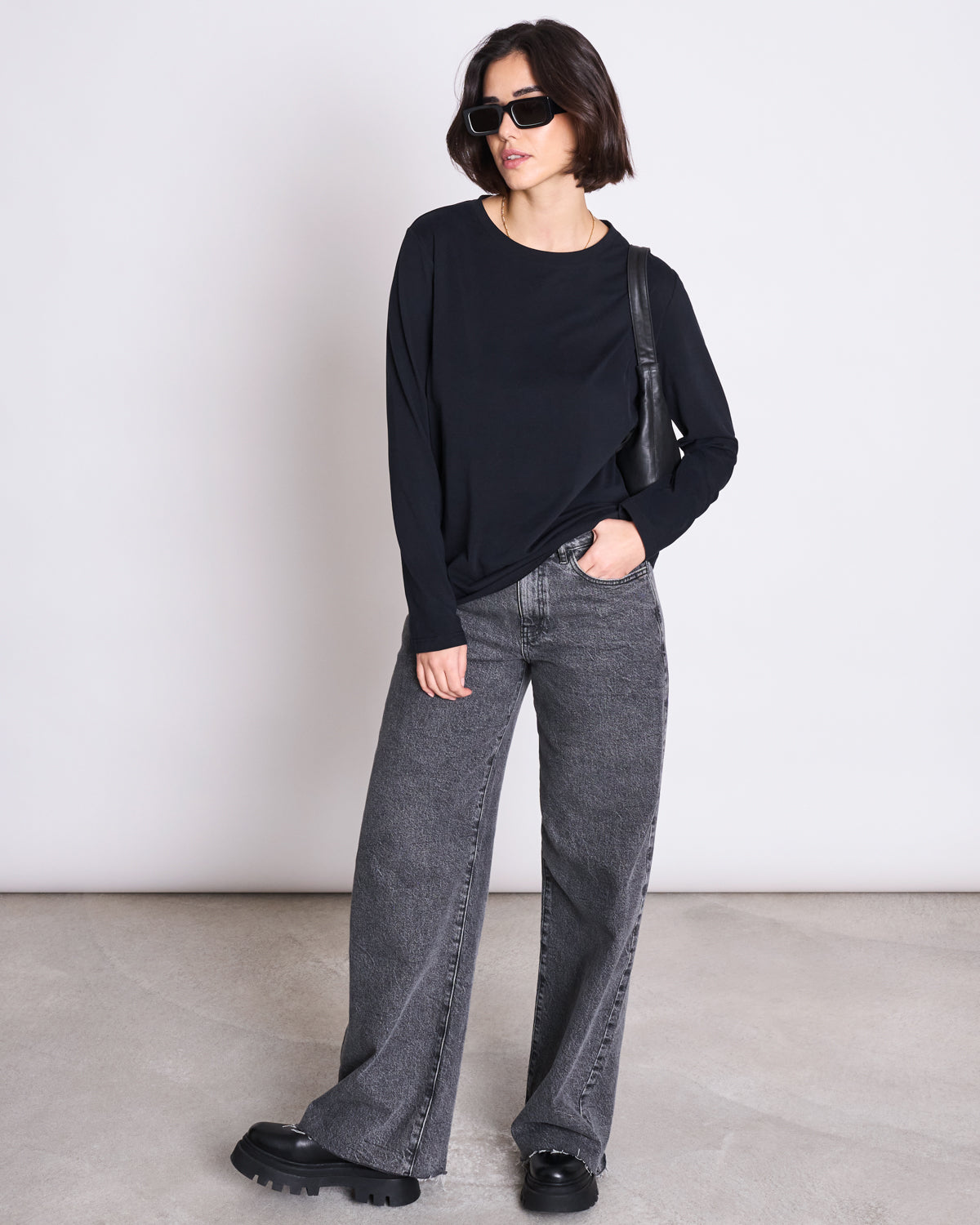 A woman wears jan-n-june’s BRUSHED LONGSLEEVE BOY BLACK GOTS top with black sunglasses, gray wide-leg jeans, and black chunky shoes, standing against a plain light background with one hand in her pocket.