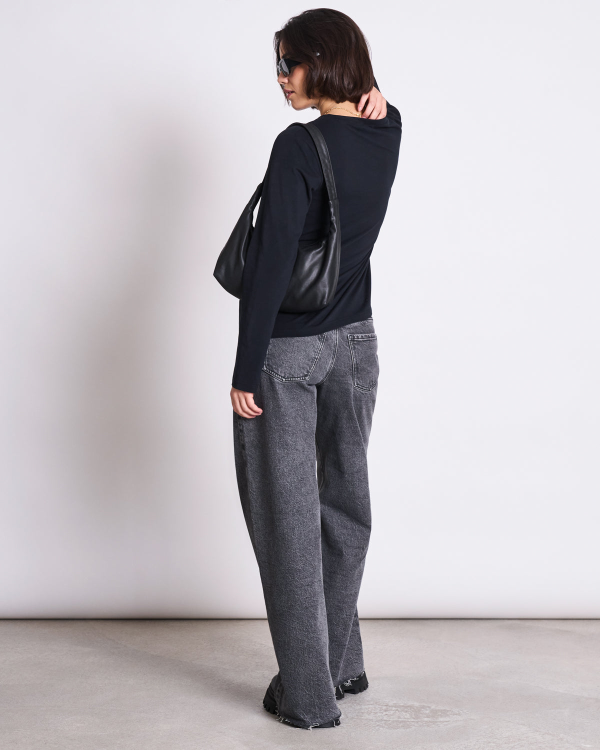 A person with short brown hair and sunglasses models jan-n-june’s BRUSHED LONGSLEEVE BOY BLACK GOTS top, paired with gray loose jeans and black shoes, standing with a black shoulder bag against a plain light background.