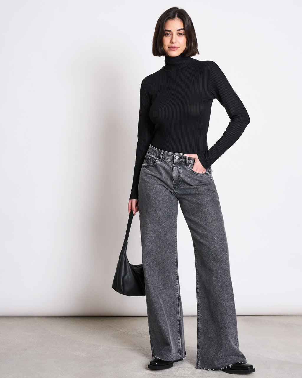 A woman poses confidently against a plain background wearing the jan-n-june LYOCELL RIB TURTLENECK MIO BLACK, paired with gray wide-leg jeans, black shoes, and holding a black handbag.