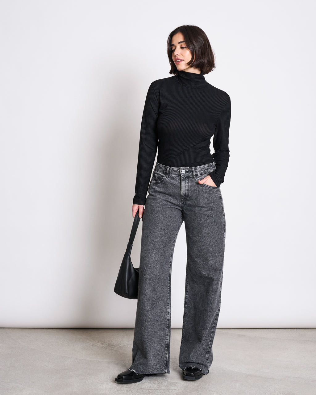 A woman poses against a plain background wearing the jan-n-june LYOCELL RIB TURTLENECK MIO BLACK, loose gray jeans, and black shoes. She holds a black handbag, looking to the side with one hand in her pocket.