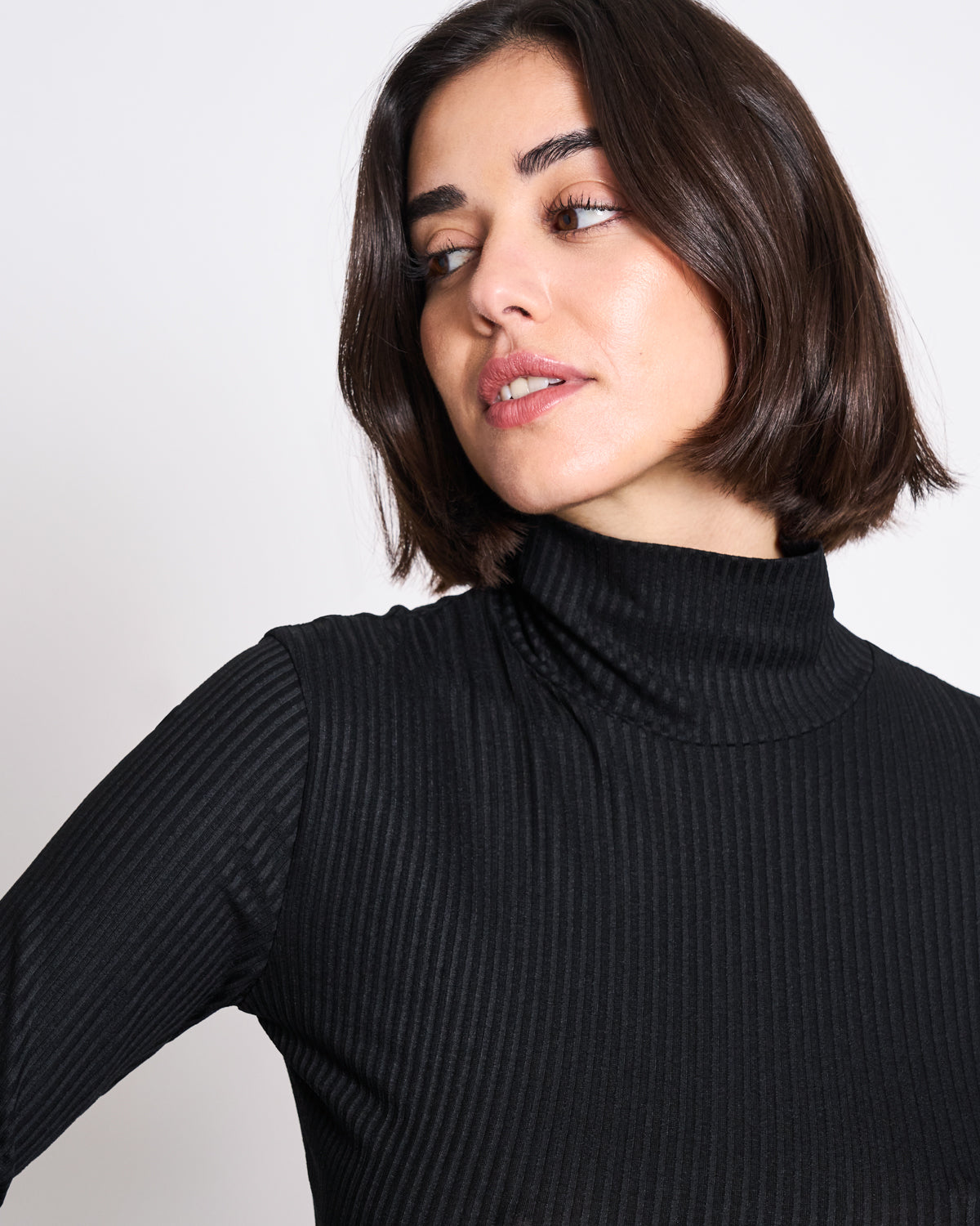 A woman with chin-length dark hair wears the jan-n-june LYOCELL RIB TURTLENECK MIO BLACK, looking to her left against a plain white background.