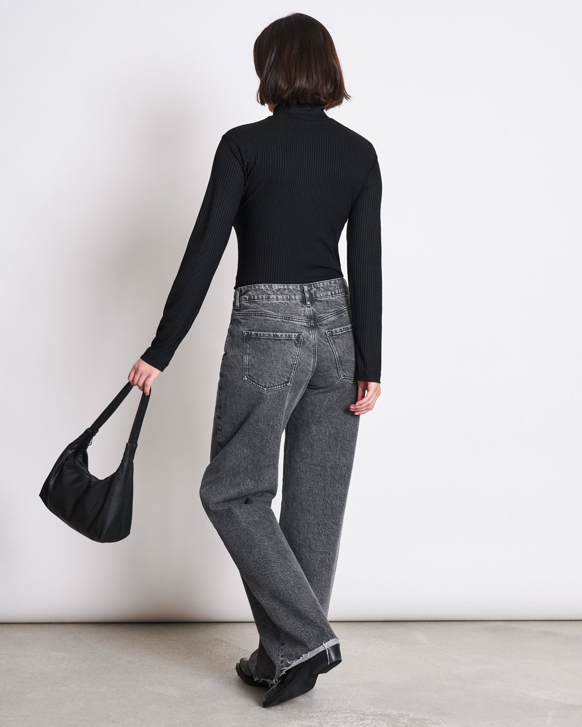 A person with short dark hair, wearing the jan-n-june LYOCELL RIB TURTLENECK MIO BLACK, gray high-waisted jeans, and black shoes stands facing away against a plain wall, holding a small black handbag in their left hand.