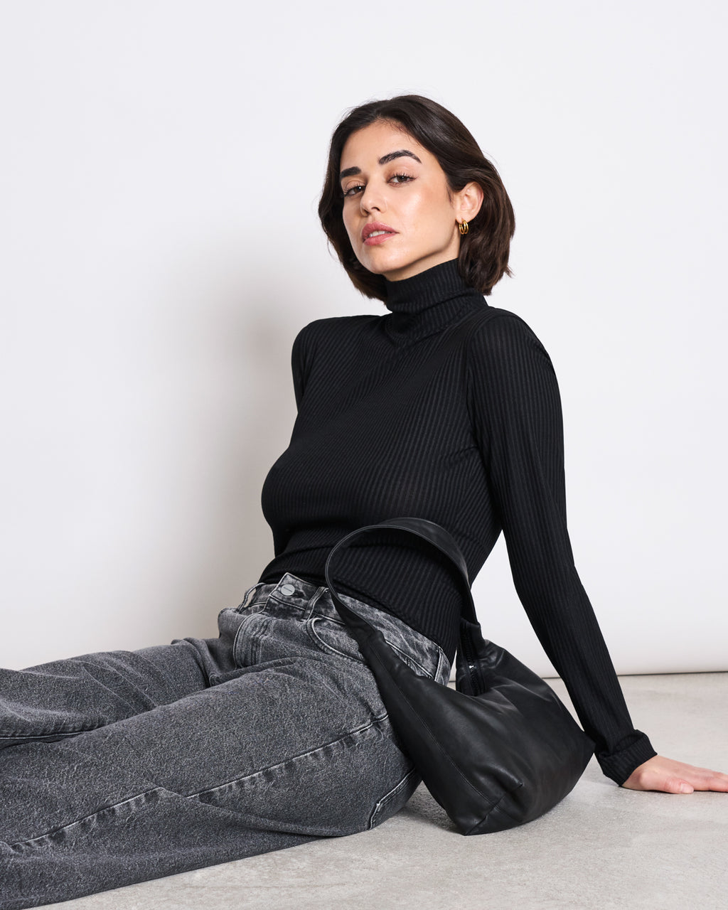 A woman with short dark hair wears the jan-n-june LYOCELL RIB TURTLENECK MIO BLACK and gray jeans, sitting on the floor against a white background with a black handbag by her side.