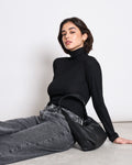 A woman with short dark hair wears the jan-n-june LYOCELL RIB TURTLENECK MIO BLACK and gray jeans, sitting on the floor against a white background with a black handbag by her side.