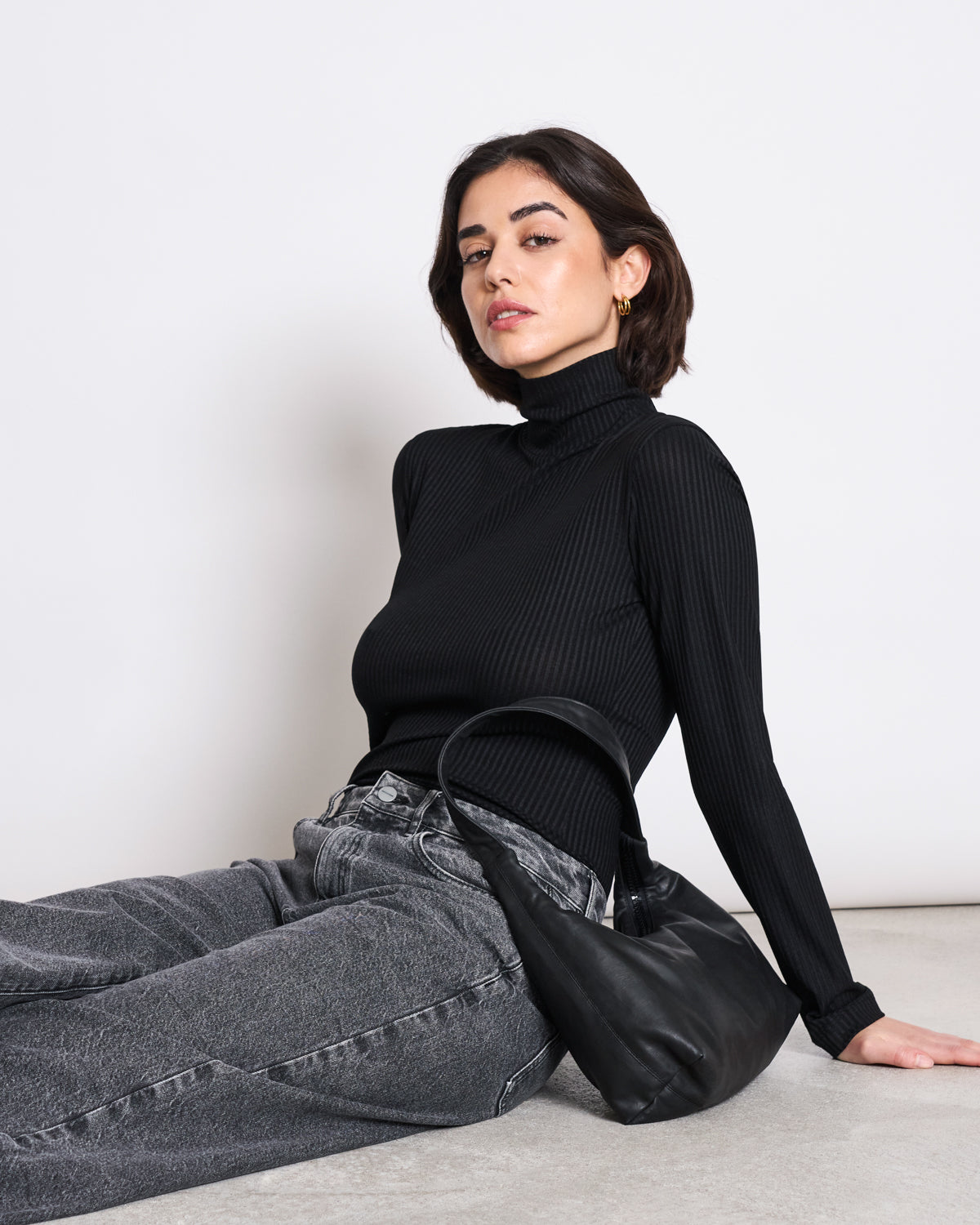 A woman with short dark hair wears the jan-n-june LYOCELL RIB TURTLENECK MIO BLACK and gray jeans, sitting on the floor against a white background with a black handbag by her side.