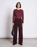 A woman with a short dark bob faces forward, wearing the jan-n-june RIB LONGSLEEVE MURI INTENSE RUST GOTS top and matching high-waisted wide-leg trousers. She holds a black handbag and wears black pointed shoes against a plain white background.
