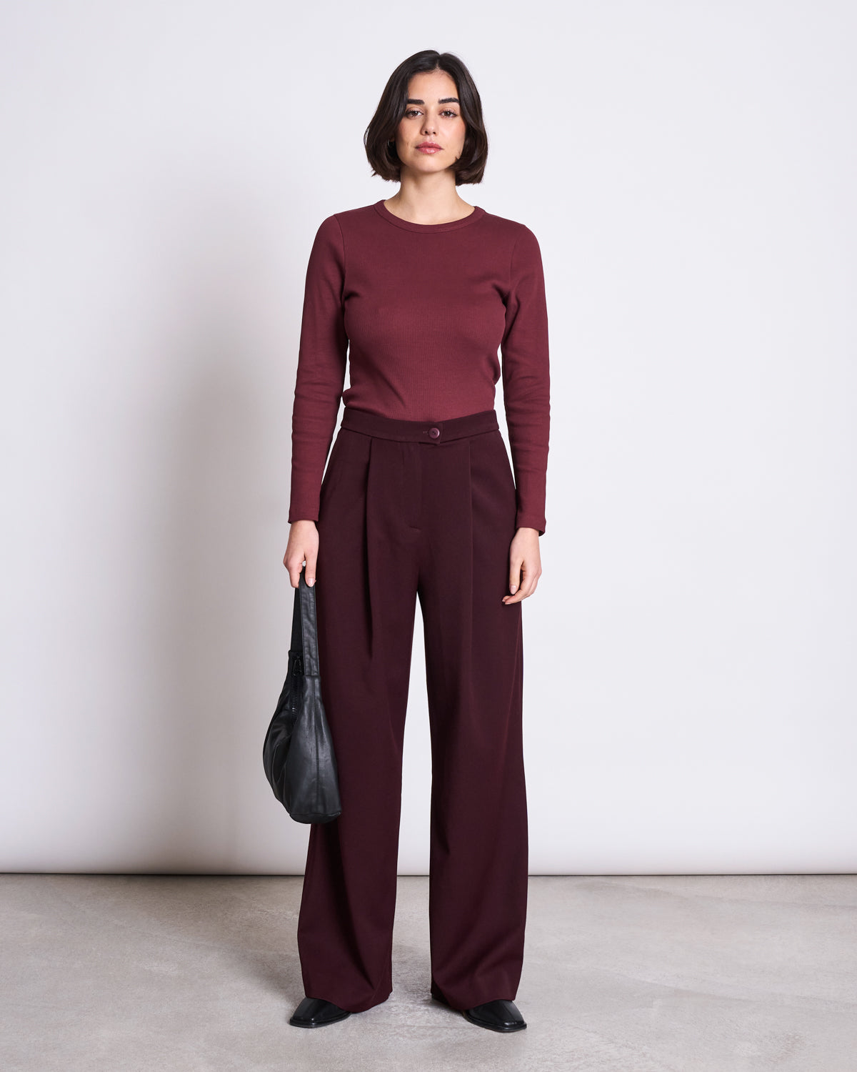 A woman with a short dark bob faces forward, wearing the jan-n-june RIB LONGSLEEVE MURI INTENSE RUST GOTS top and matching high-waisted wide-leg trousers. She holds a black handbag and wears black pointed shoes against a plain white background.