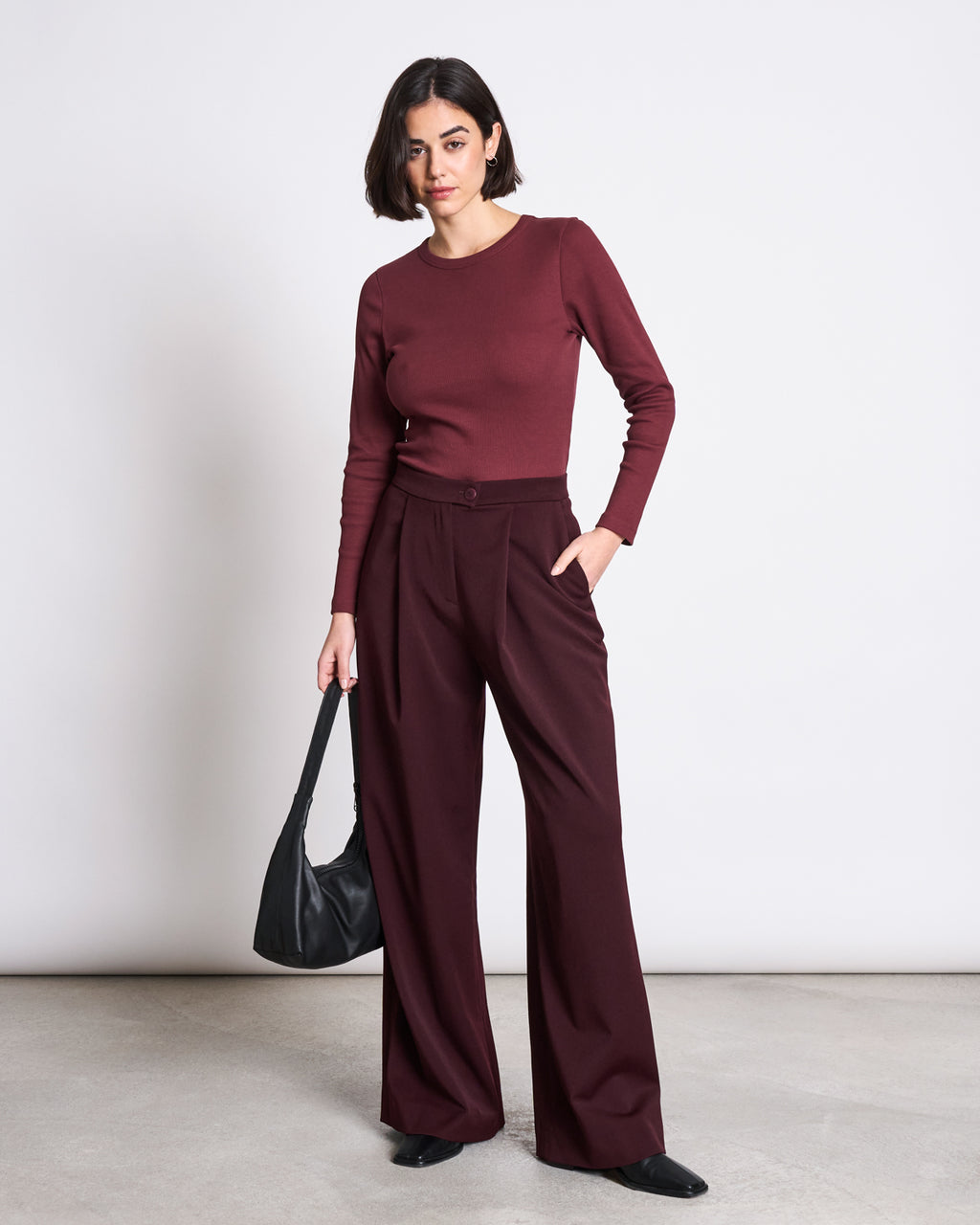 A woman stands against a plain background wearing jan-n-junes RIB LONGSLEEVE MURI INTENSE RUST GOTS with matching wide-leg pants, holding a black shoulder bag and wearing black shoes. Her short dark hair is simply styled—a timeless wardrobe essential.