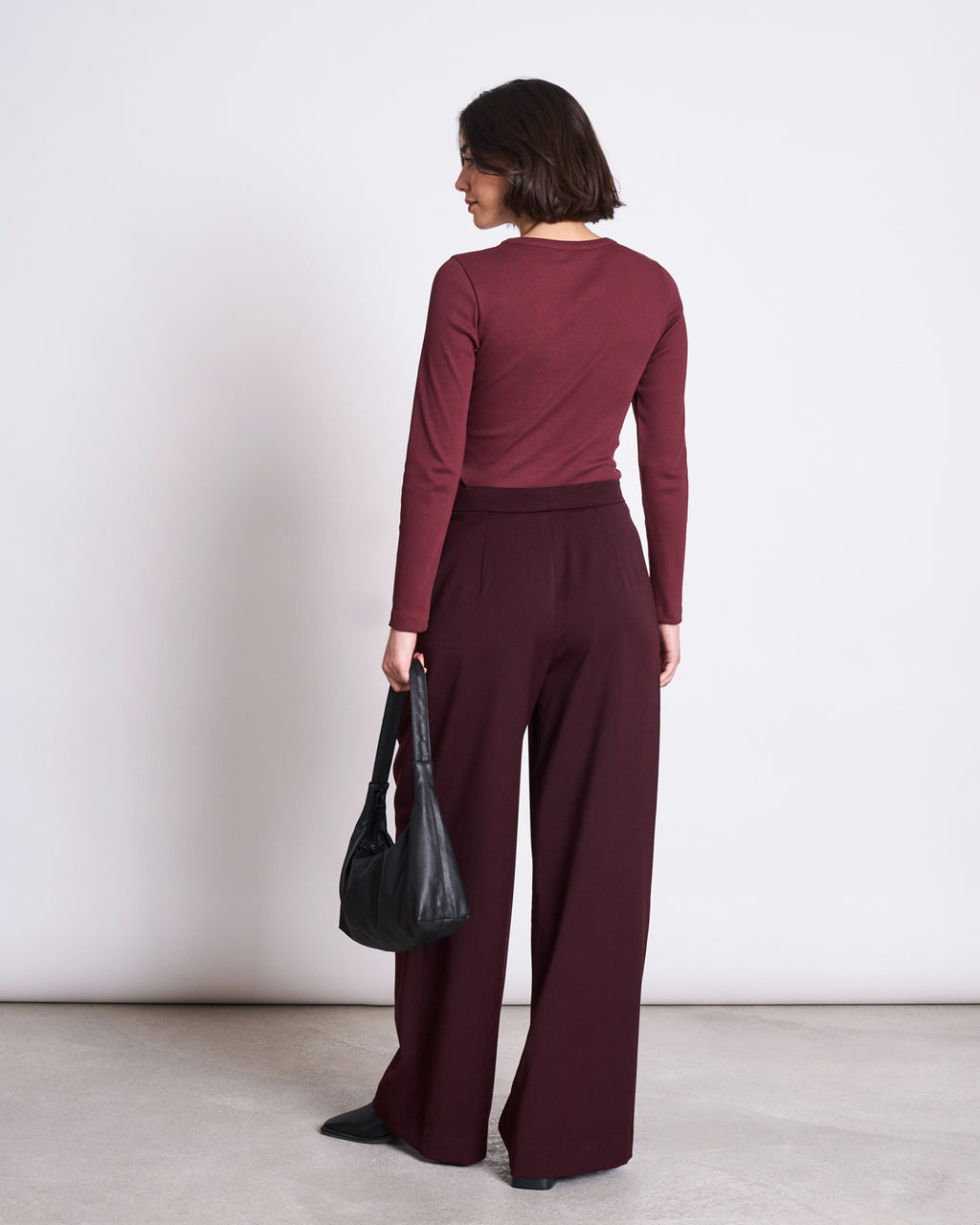 A woman faces away in the jan-n-june RIB LONGSLEEVE MURI INTENSE RUST GOTS, paired with wide-leg burgundy pants. She holds a black handbag and stands on a light floor against a plain white background.