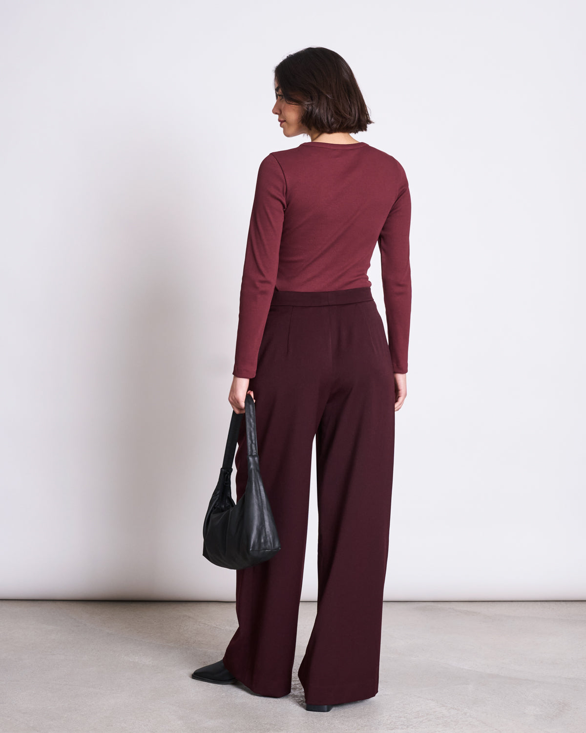 A woman faces away in the jan-n-june RIB LONGSLEEVE MURI INTENSE RUST GOTS, paired with wide-leg burgundy pants. She holds a black handbag and stands on a light floor against a plain white background.
