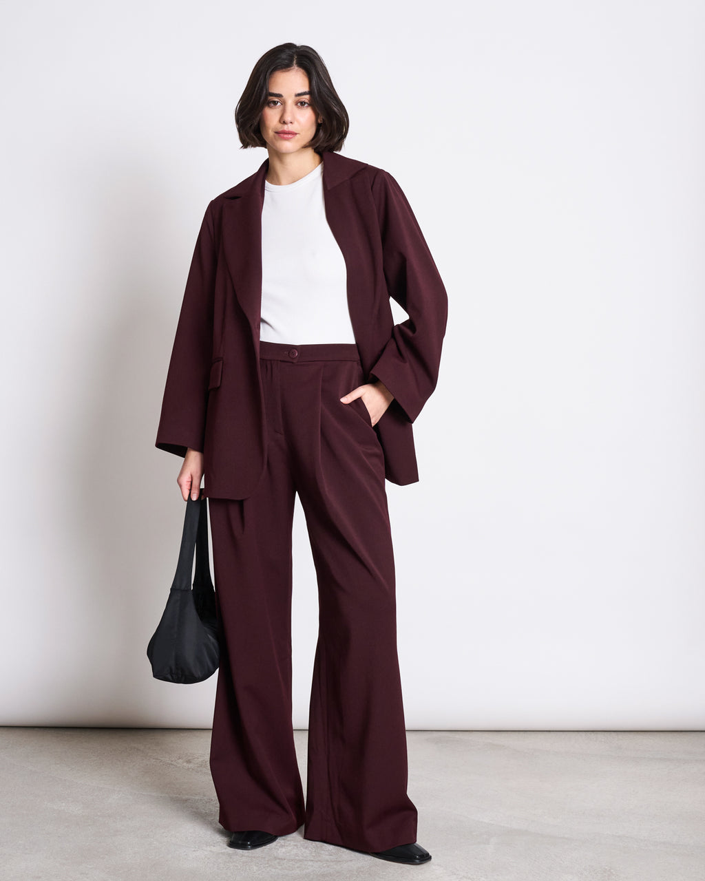 A woman with short dark hair wears the jan-n-june BLAZER DURANCE INTENSE RUST, made from recycled polyester, over a white top with black shoes. She stands against a plain background, holding a black shoulder bag and looking confidently at the camera.