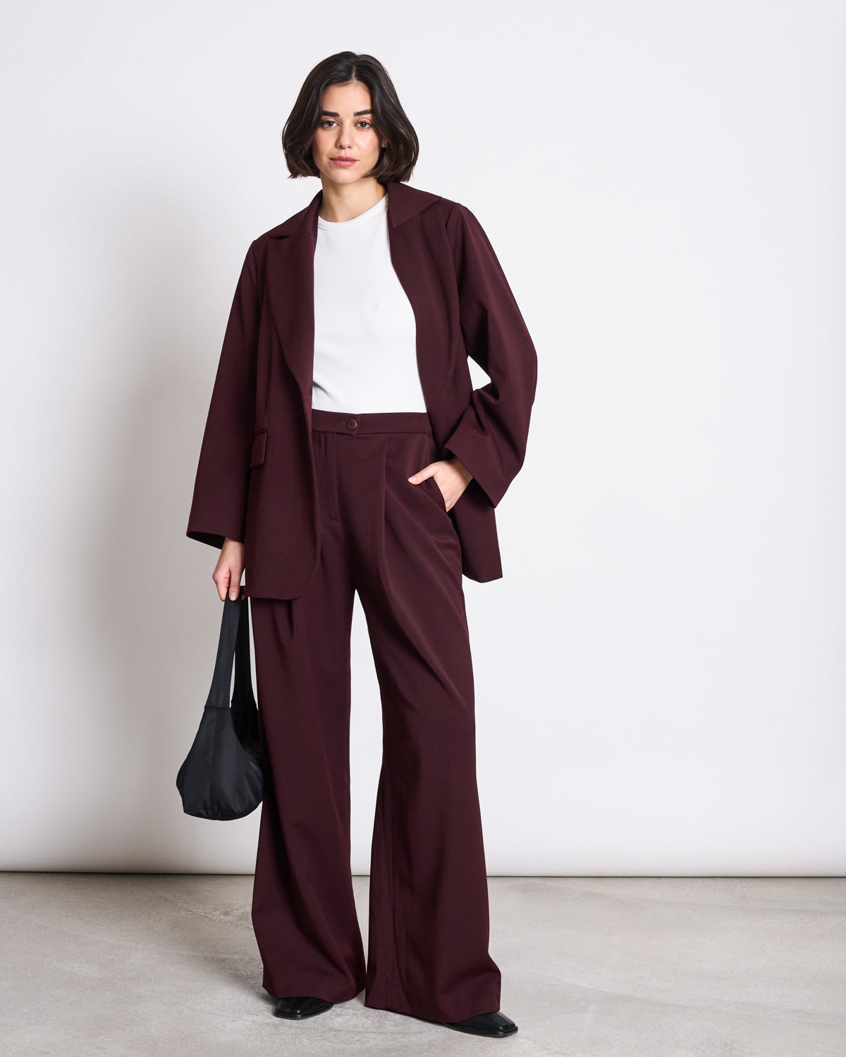 A woman with short dark hair wears the jan-n-june BLAZER DURANCE INTENSE RUST, made from recycled polyester, over a white top with black shoes. She stands against a plain background, holding a black shoulder bag and looking confidently at the camera.