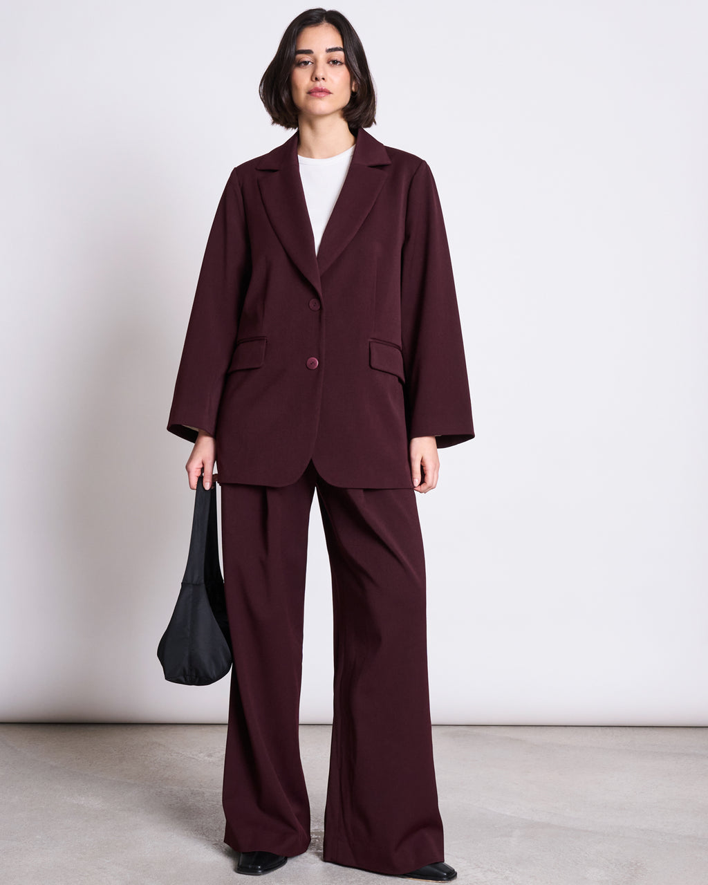 A person stands against a plain background wearing the jan-n-june BLAZER DURANCE INTENSE RUST—an oversized, burgundy blazer made from recycled polyester—paired with a white top, black shoes, and holding a black handbag.
