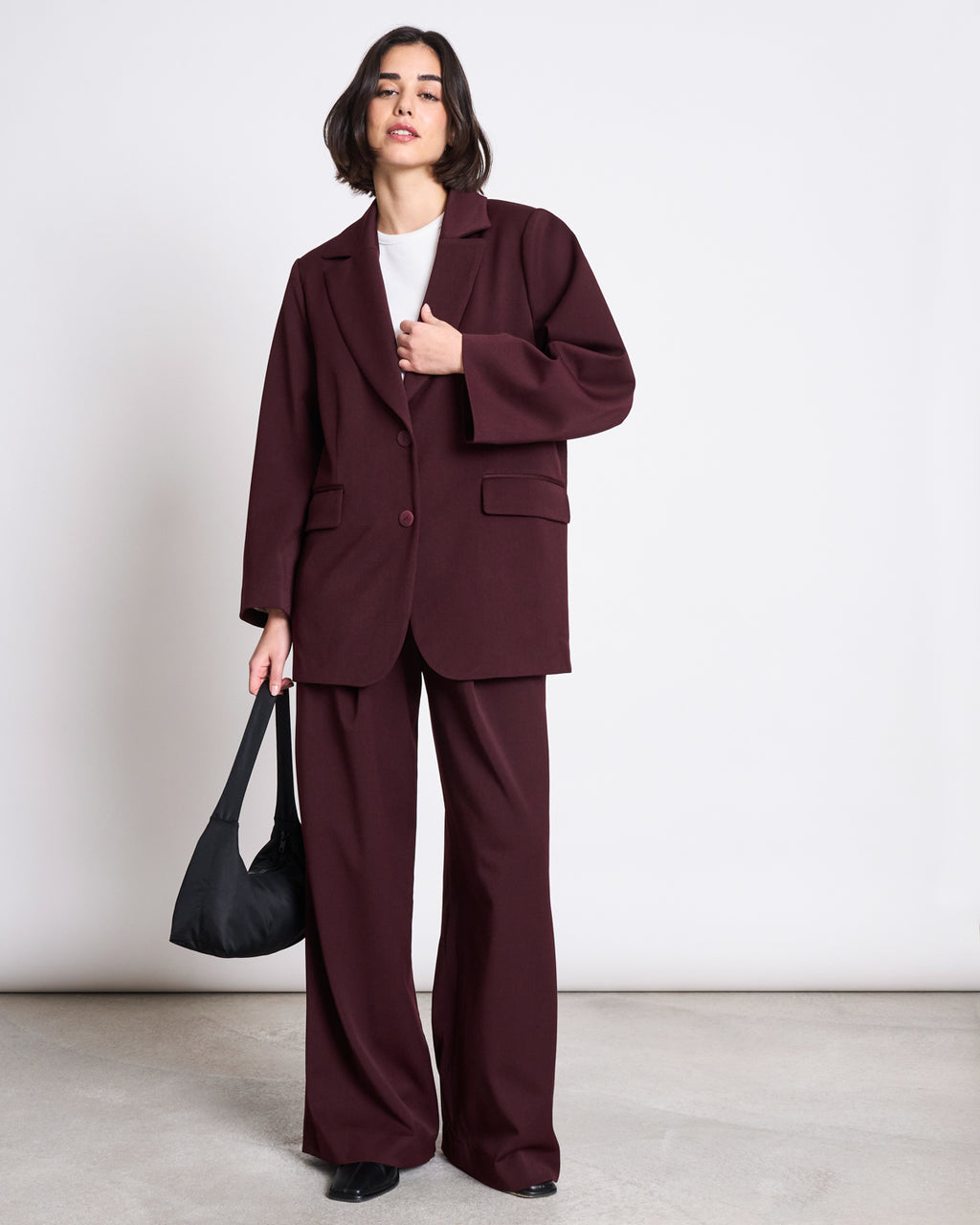 A woman with short dark hair wears the jan-n-june BLAZER DURANCE INTENSE RUST suit—an oversized burgundy blazer with wide-leg pants over a white top, carrying a black handbag, standing against a plain white background.