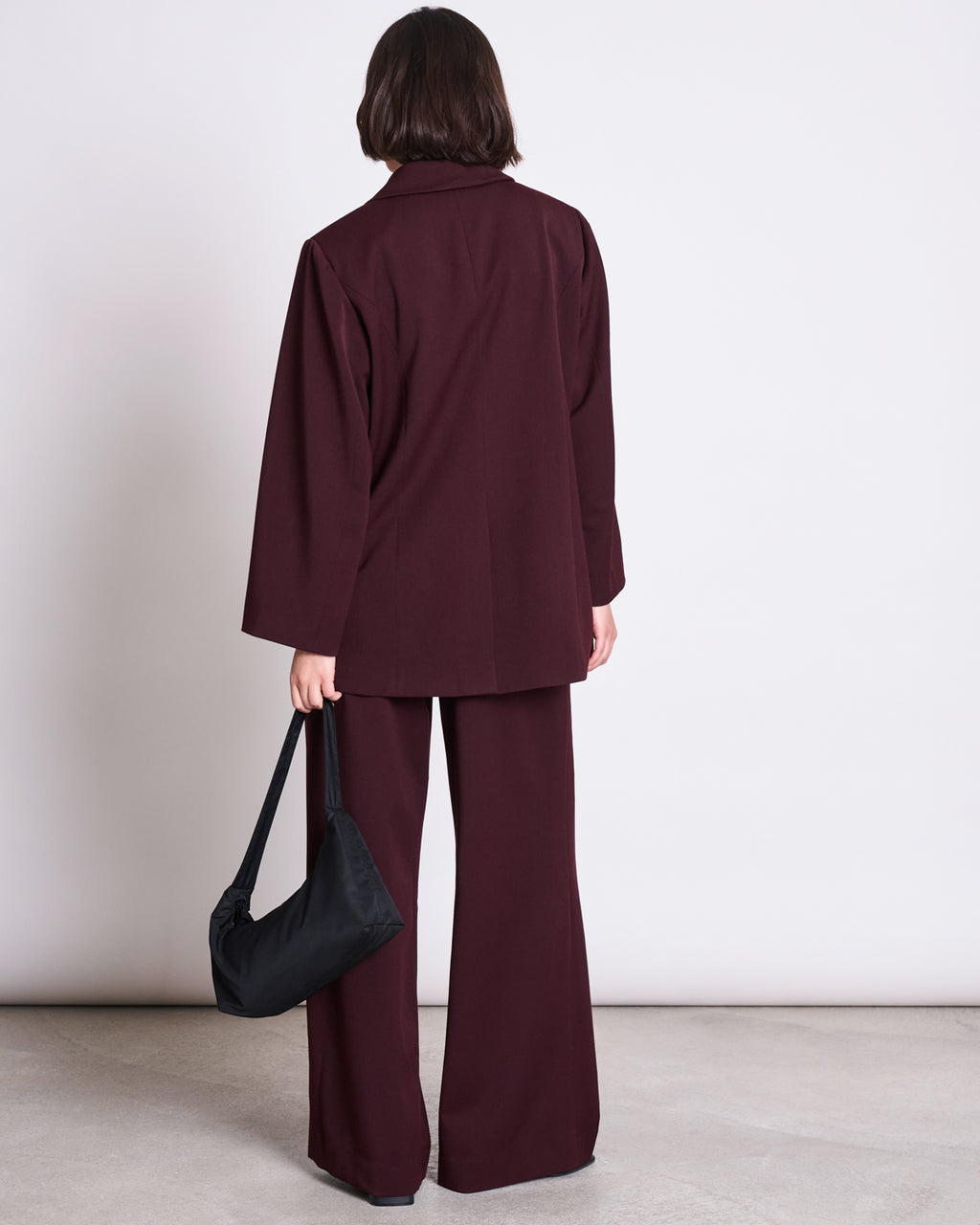 A person with short dark hair, seen from behind, wears the jan-n-june BLAZER DURANCE INTENSE RUST with wide-leg trousers and holds a black shoulder bag, standing against a plain light background.