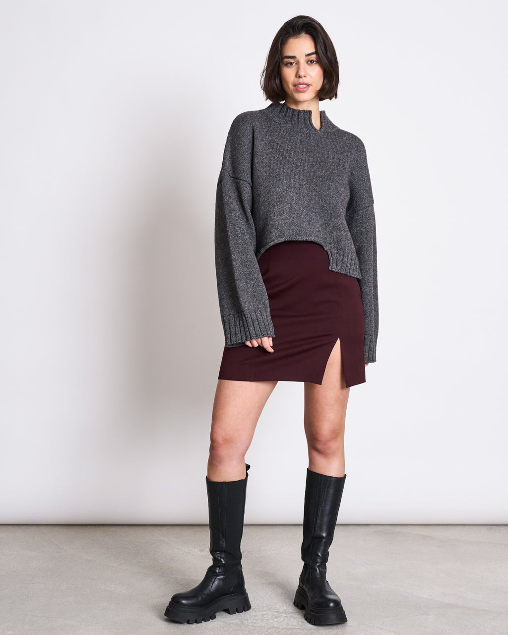 A woman with short dark hair and a confident expression wears a gray EcoVero™ oversized sweater, the jan-n-june MINI SKIRT AINOHA INTENSE RUST with a thigh slit, and black knee-high boots against a plain background.