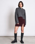 A woman with short dark hair and a confident expression wears a gray EcoVero™ oversized sweater, the jan-n-june MINI SKIRT AINOHA INTENSE RUST with a thigh slit, and black knee-high boots against a plain background.
